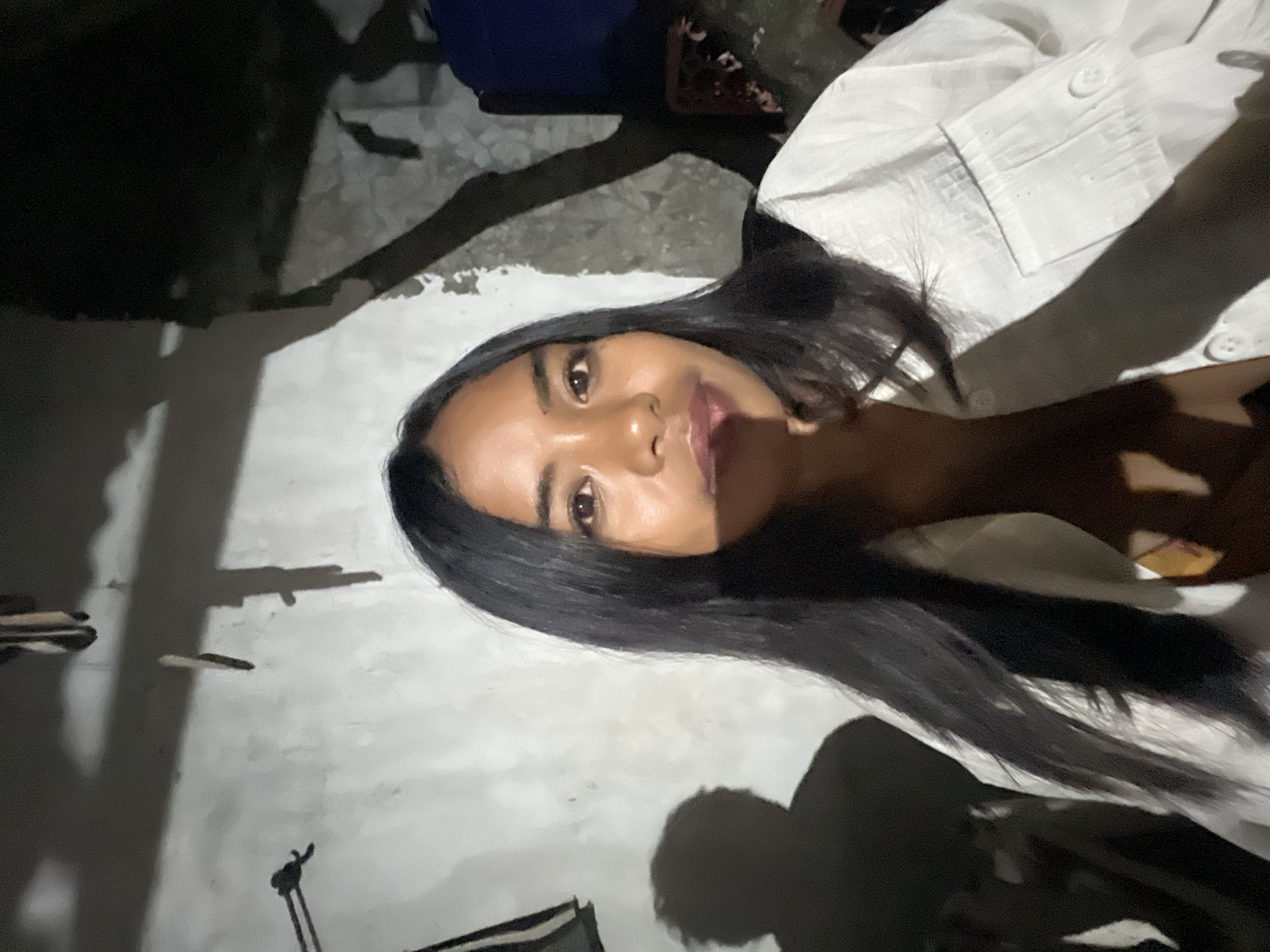 A young woman with long dark hair is captured in a close-up, selfie-style shot against a textured, light-colored wall. She is wearing a white button-up shirt. The lighting is dim, creating strong shadows on the wall, including the shadow of the woman and what appears to be a tree branch or structure. The background is dimly lit, with some indistinct objects and the shadow of a person on the right side. The overall impression is that of an outdoor setting at night, possibly in a residential area of Denpasar, Indonesia, given the architectural style of the wall and the presence of vegetation shadows.
