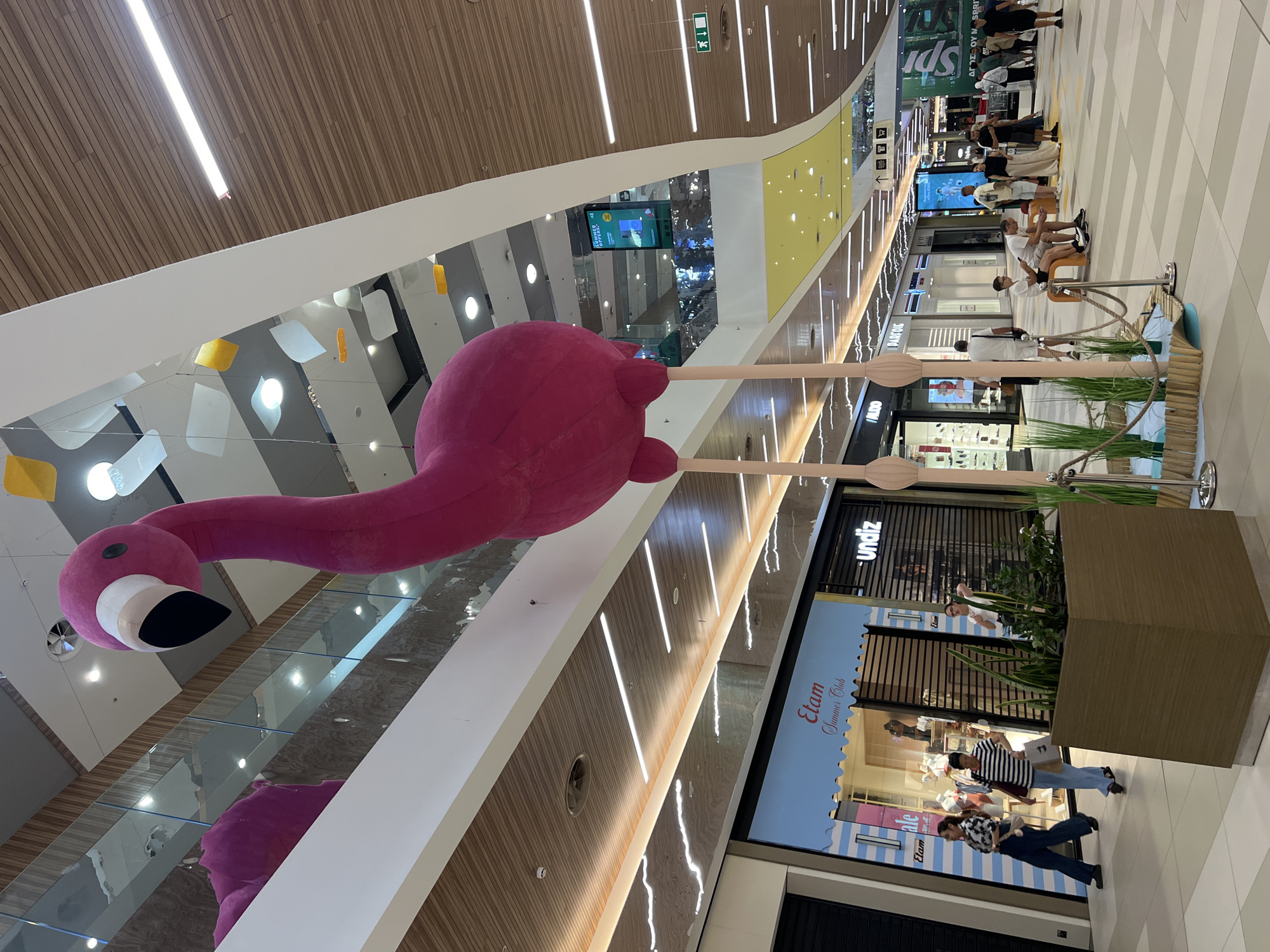 The image shows a large, inflatable pink flamingo sculpture suspended from the ceiling of a modern indoor shopping mall. The flamingo has a long neck, a black beak, and stands on two thin, pink legs. It is positioned in the foreground, dominating the center of the frame. The mall environment features multiple levels, with balconies and walkways visible above. The ceiling is a combination of wooden panels and white structures with integrated lighting. Below the flamingo, a planter with green foliage and decorative blue water elements is visible. In the background, various retail stores with illuminated signage are present, including "Etam," "undiz," and "ALDO." Several people are walking and standing in the mall, engaged in shopping or leisure activities. The overall scene depicts a vibrant and contemporary commercial space. This mall is located in Lakatameia, Cyprus.