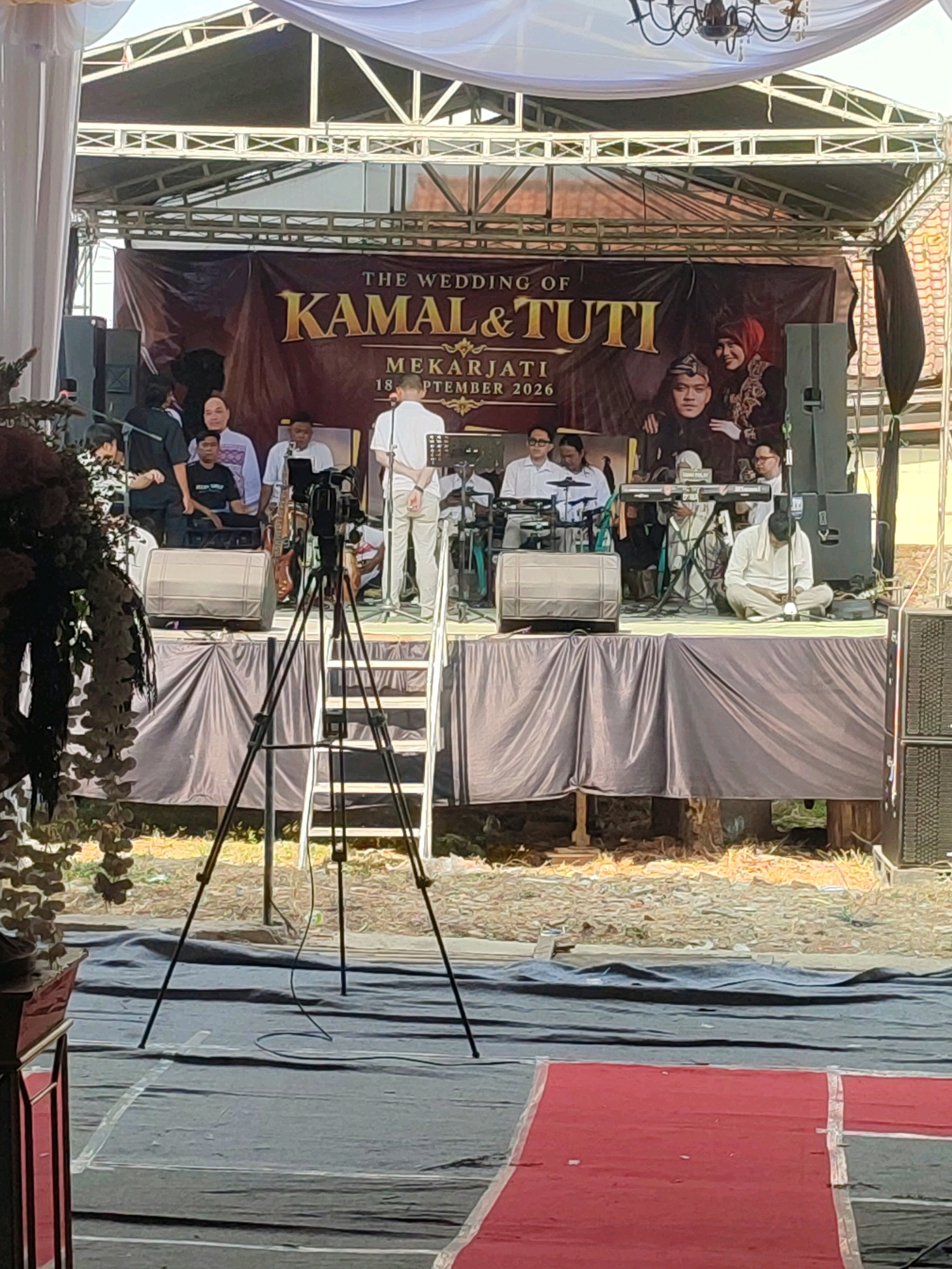 This image shows an outdoor wedding reception stage set up in Kecamatan Haurgeulis, Indonesia. A large banner behind the performers reads "THE WEDDING OF KAMAL & TUTI MEKARJATI 18 SEPTEMBER 2026." A band is performing on the stage, with several musicians playing instruments and a person singing. In the foreground, a video camera on a tripod is positioned to record the event. The stage is draped with dark fabric, and speakers are set up on either side. The ground in front of the stage is covered with dark tarpaulin, with a red carpet leading towards the stage. The overall scene suggests a festive and celebratory atmosphere.