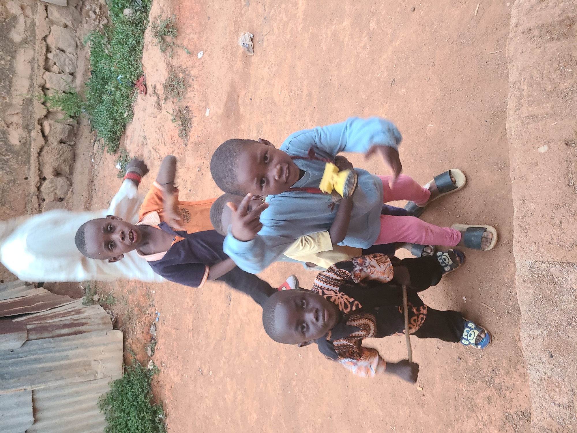 A high-angle shot depicts four children in Jos, Nigeria, posing together outdoors on a dirt ground with patches of vegetation. The child in the foreground is wearing a dark, patterned top and black pants with blue and white patterned sandals. The second child from the foreground wears a yellow shirt. Above that child, a third child is wearing a light blue top and pink pants with black sandals. The fourth and furthest child wears an orange shirt. In the background, there is a stone wall and a corrugated metal structure.