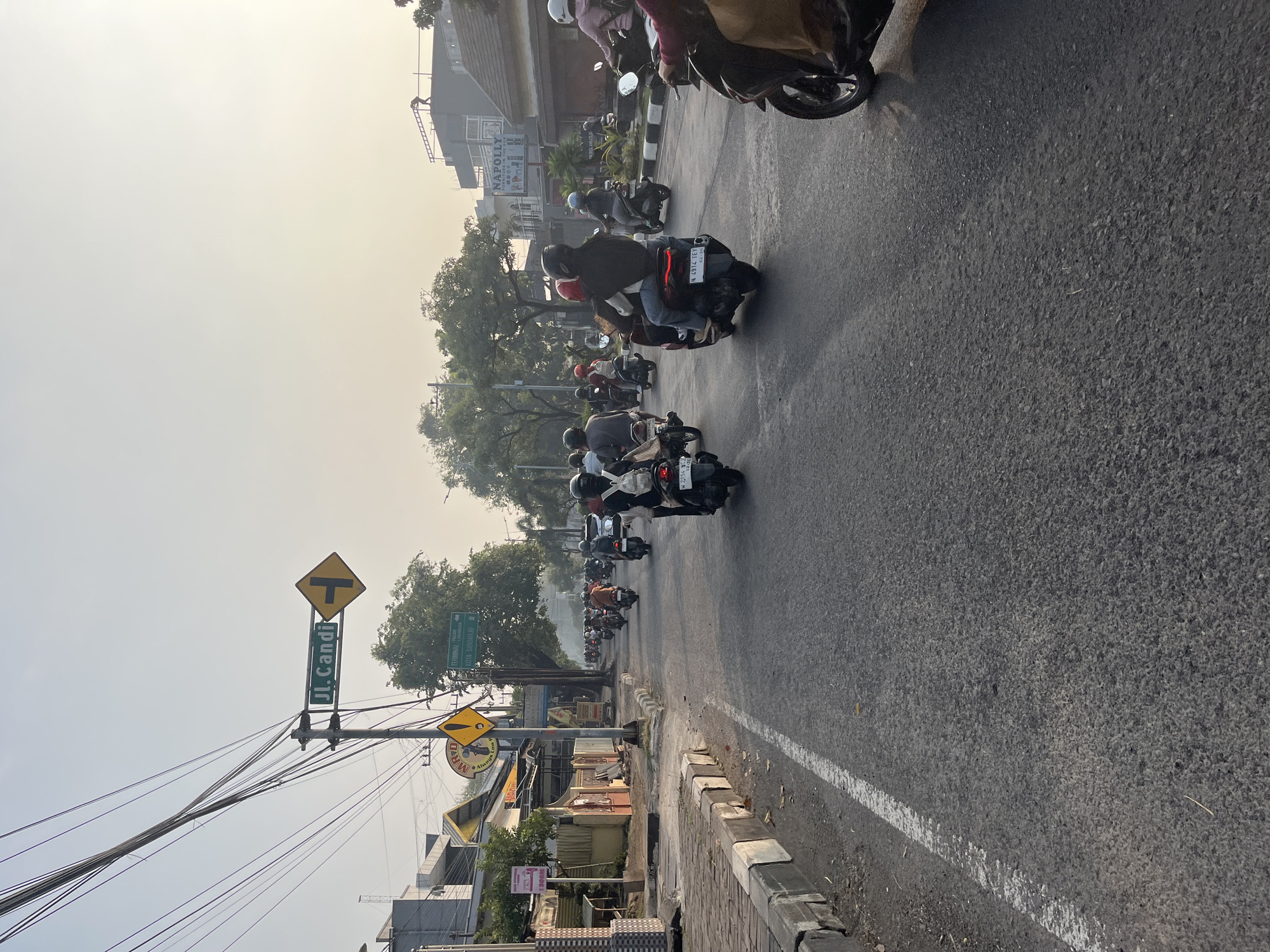 The image depicts a street scene in Tenggulunan, Indonesia, likely during the daytime with hazy or overcast skies. A stream of motorcycles is traveling along a paved road. Many of the riders are wearing helmets and some have passengers. On the left side of the road, there is a sidewalk with a curb and various businesses and structures. A street sign indicates "Jl. Candi" and a directional arrow. Another yellow diamond-shaped sign with an exclamation mark is visible. Trees and buildings line the street. The overall atmosphere suggests a busy urban environment with moderate traffic.