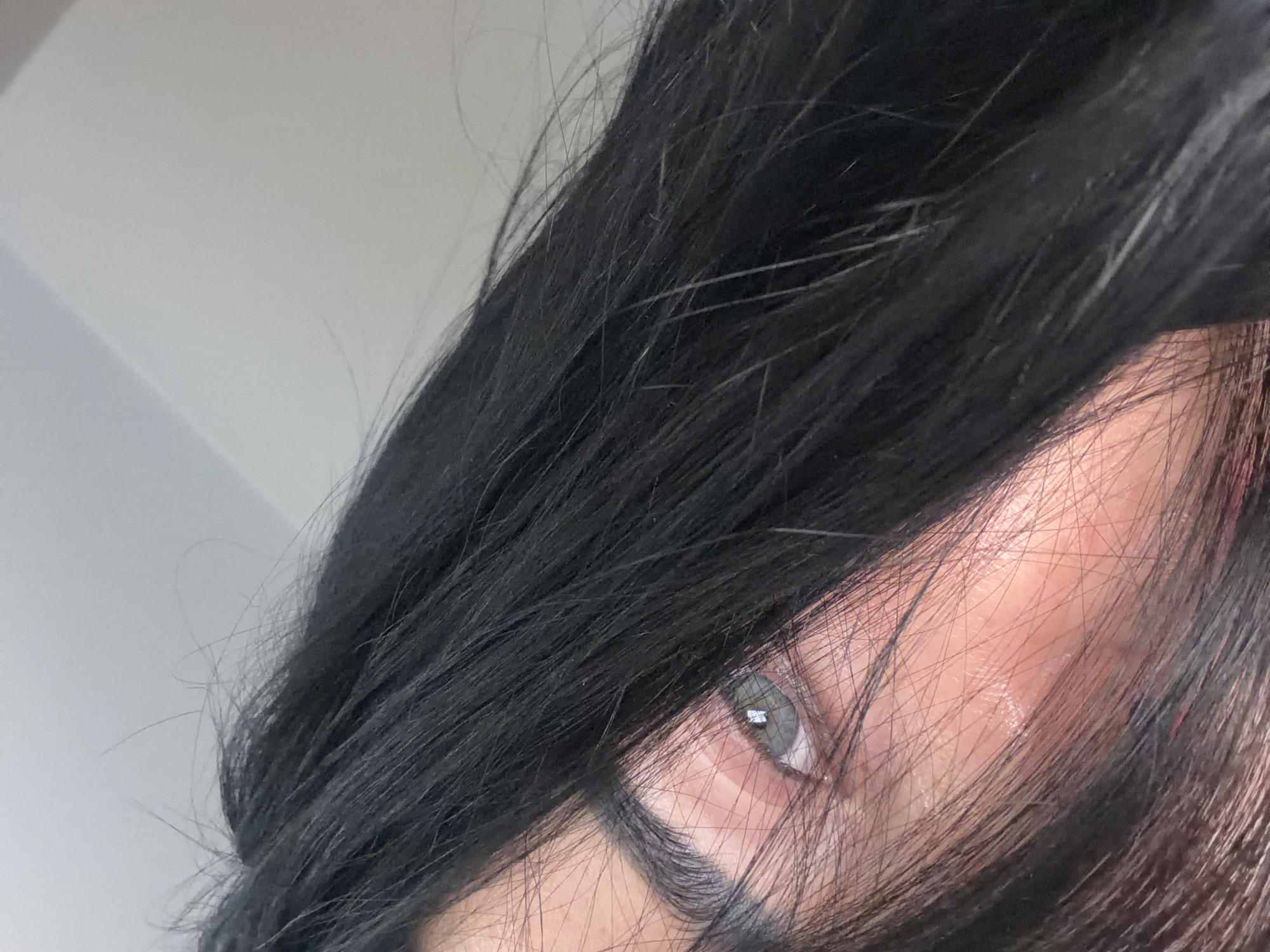 The image is a close-up shot featuring a person's face partially obscured by dark hair. One eye is visible, appearing gray or light-colored. The skin is pale and reflects light. The background is a plain, light gray or white, suggesting an indoor setting. There is no indication of Saint Petersburg, Russia, or any city.