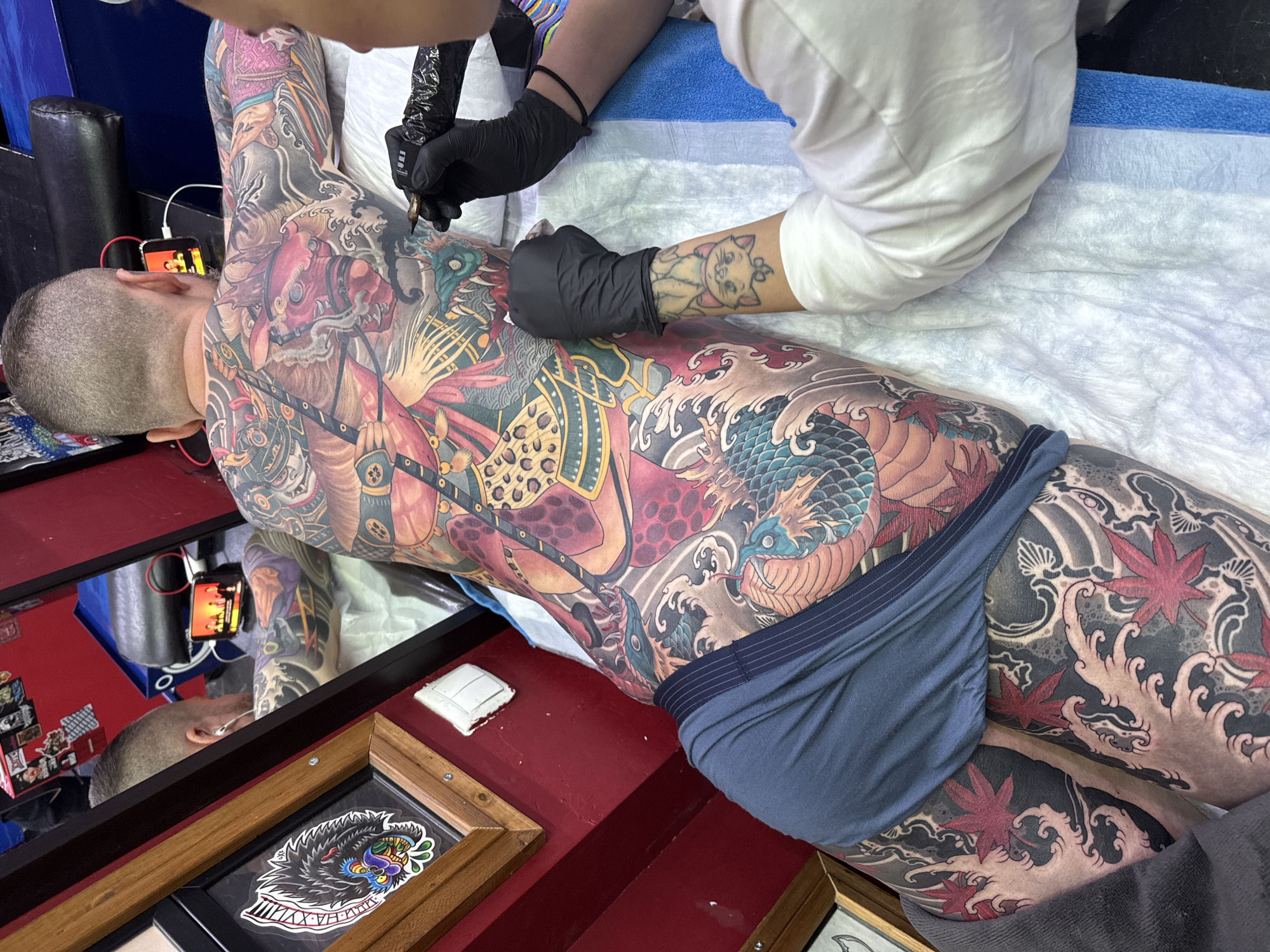 A person is receiving a full-body tattoo in what appears to be a tattoo parlor. The individual is lying face down on a white sheet, wearing dark blue underwear. The artist, clad in a white long-sleeved shirt and black gloves, is using a tattoo machine on the person's back. The person receiving the tattoo already has extensive tattoos covering their entire back and arms, featuring traditional Japanese designs such as dragons, waves, and maple leaves. A phone is connected to headphones is visible in the upper left of the frame. A red surface with framed tattoo artwork is in the foreground. The exact location within Moscow, Russia, cannot be determined based solely on the image.