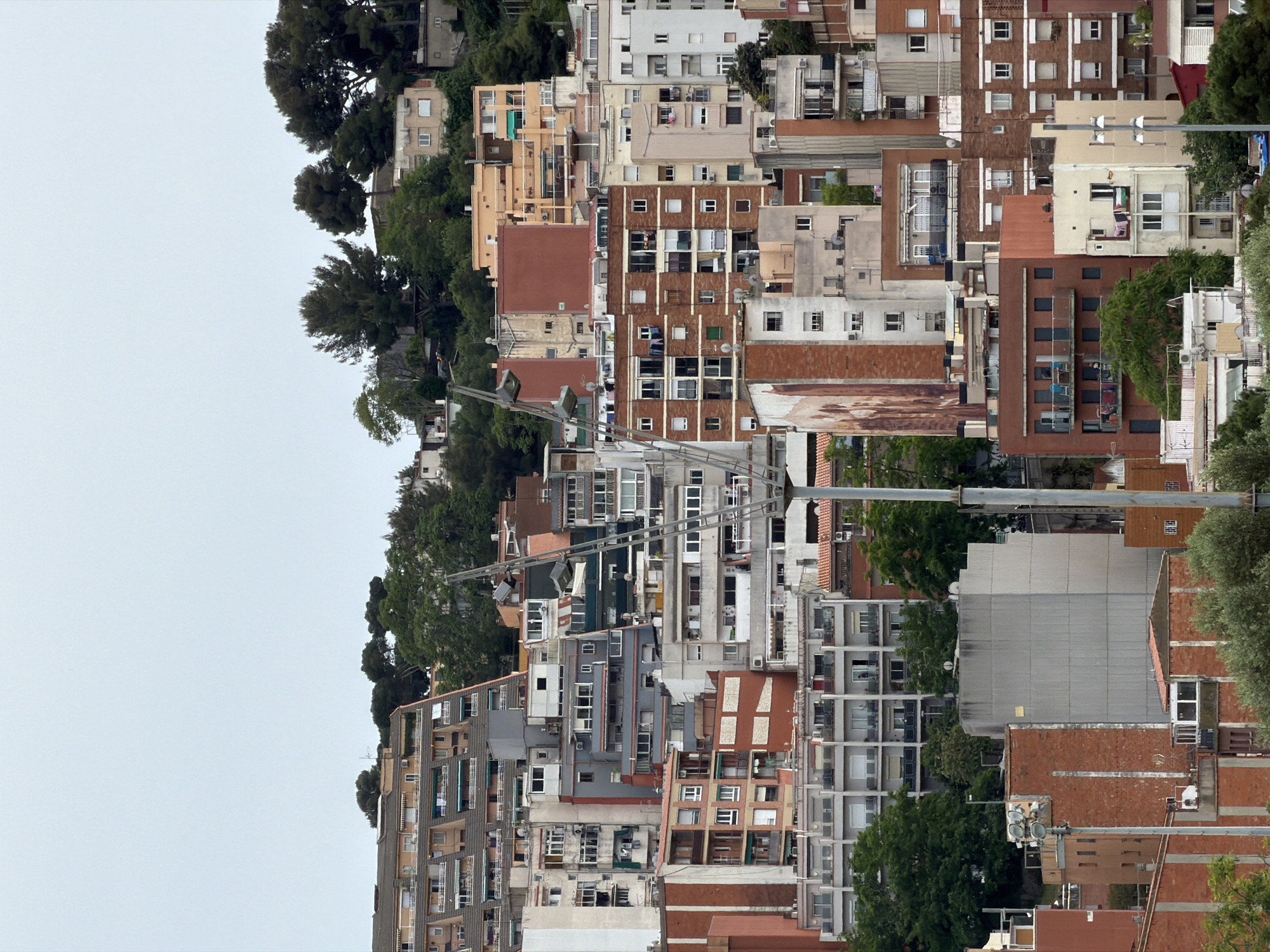 The image presents a densely packed urban scene with multiple residential buildings in what appears to be Barcelona, Spain. The architectural style is typical of Southern European urban housing, featuring a mix of materials including brick, concrete, and painted facades. The buildings vary in height and design, reflecting a heterogeneous urban development. Some buildings have balconies and windows with shutters, while others are more modern with large glass panels. Trees are interspersed among the buildings, providing pockets of green space. The overcast sky suggests a cloudy day. Utility poles and power lines are visible, indicating the infrastructure supporting the urban environment. The overall impression is of a densely populated residential area with a mix of old and new architectural styles.