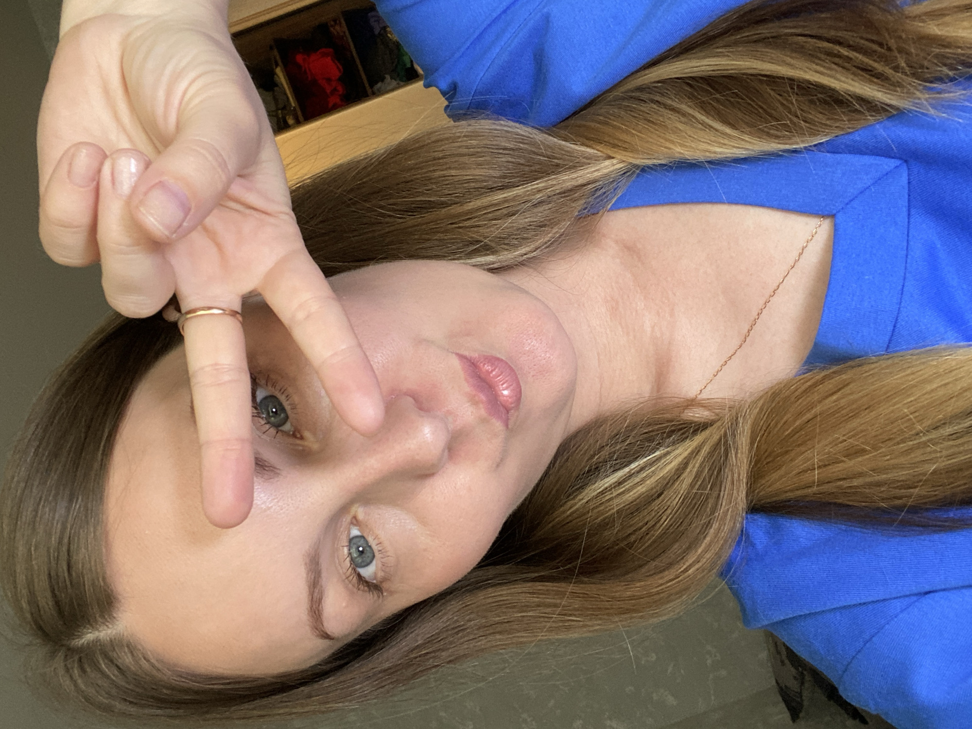 A woman with light skin and blue eyes is positioned in the center of the frame. She is holding up a hand with two fingers extended in a "peace" sign. She is wearing a blue top and a gold ring on her finger. Her hair is long and brown with blonde highlights. A gold chain is visible on her neck. A wooden cabinet with some objects inside is in the background. The image is taken at an angle, with the subject's head tilted to the side. The location of Noviy, Russia is not discernible from the image.