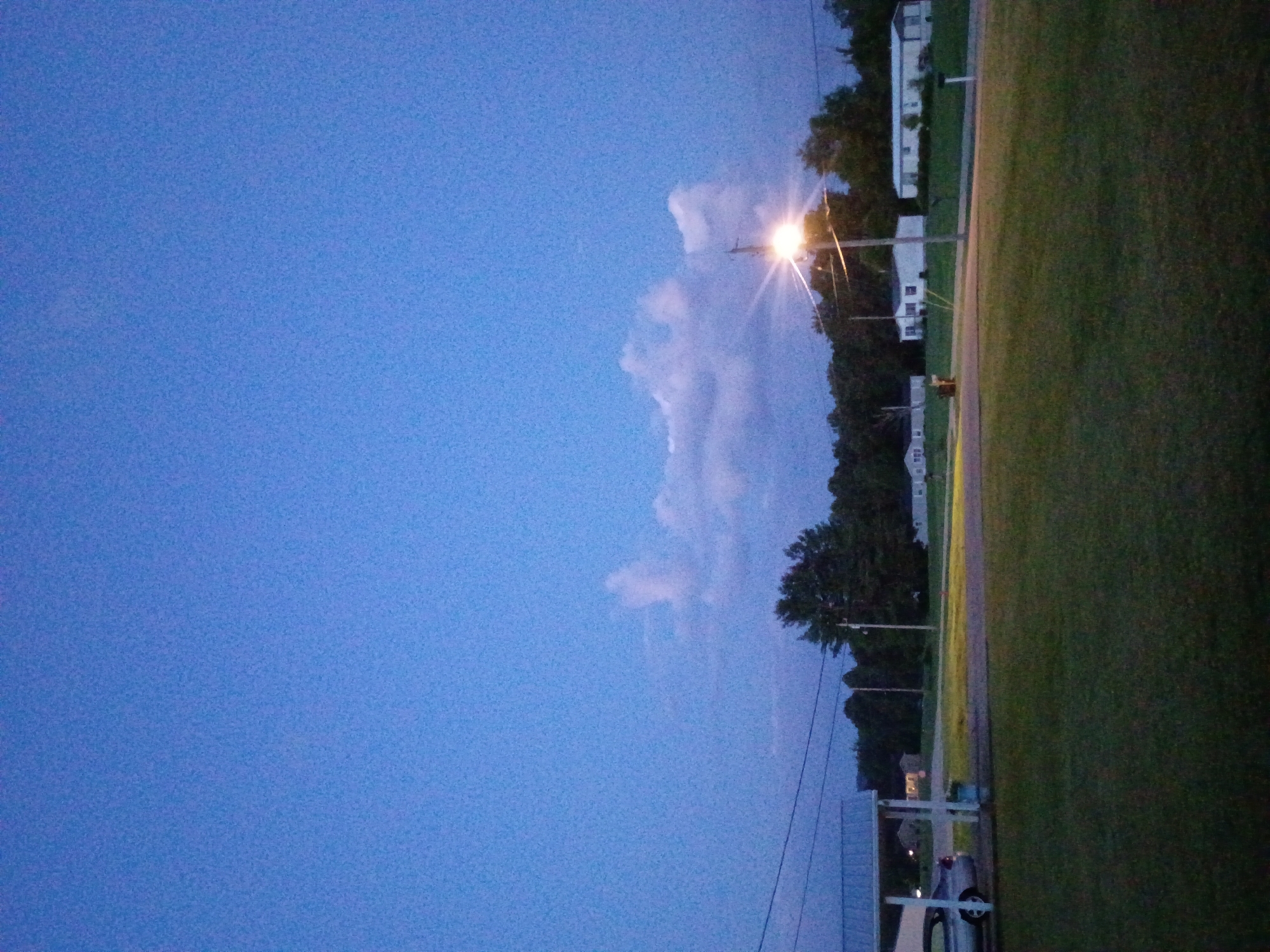 The image shows a twilight scene in a suburban area of Jeffersonville, United States of America. The sky is a deep blue, with some scattered clouds. A street light is illuminated, casting a bright glow. In the foreground, there is a grassy lawn. To the left, a silver car is parked under a carport. In the background, there are several mobile homes and trees. The overall impression is one of a quiet, peaceful evening.