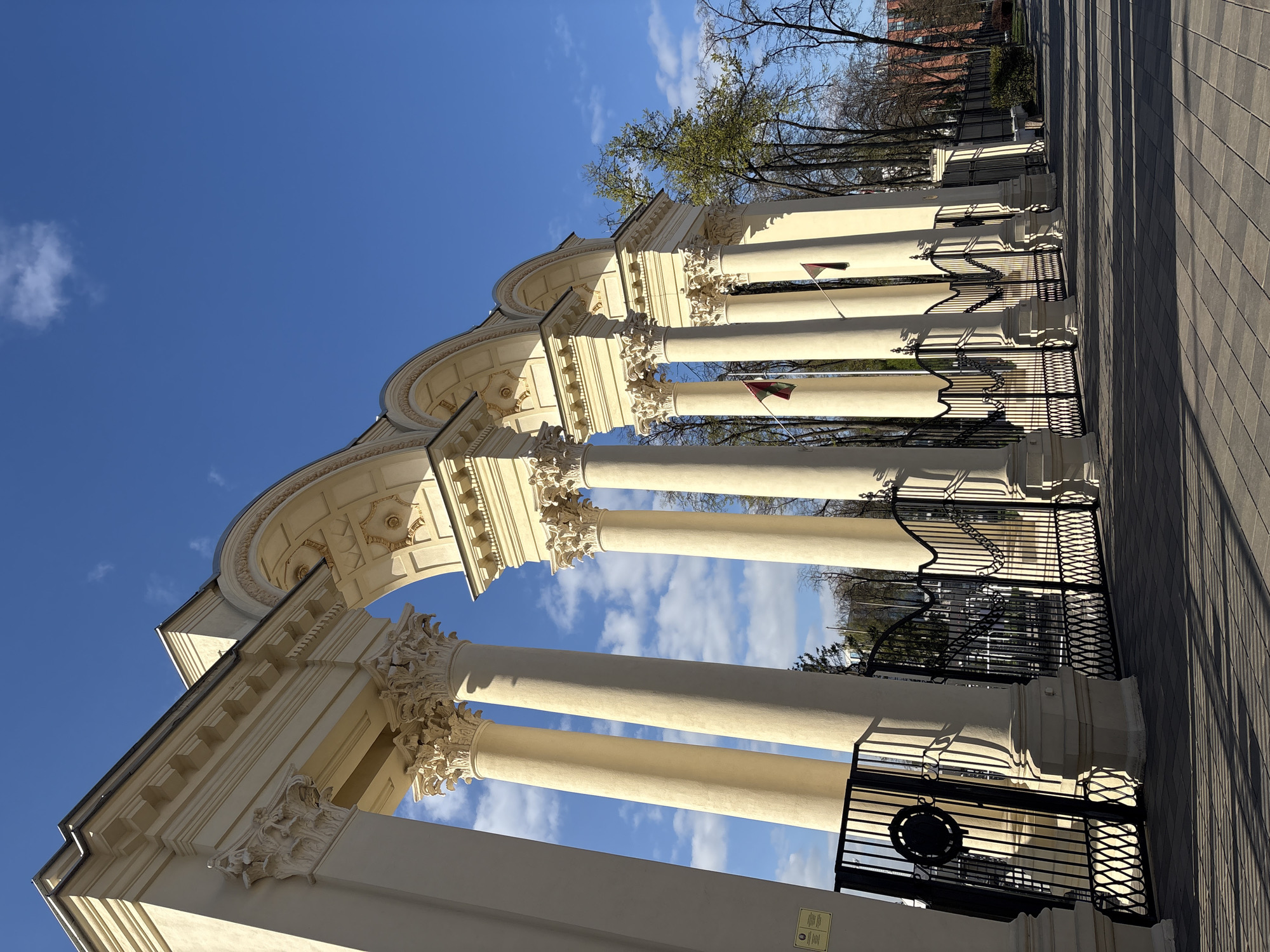The image depicts the entrance to the Gorky Park in Minsk, Belarus. The entrance is marked by a series of cream-colored classical columns topped with ornate capitals and arches, forming a colonnade. Black wrought-iron gates are interspersed between the columns. A paved pathway stretches away from the entrance. The sky is blue with scattered clouds. Some trees are visible in the background, as is a building in the distance.