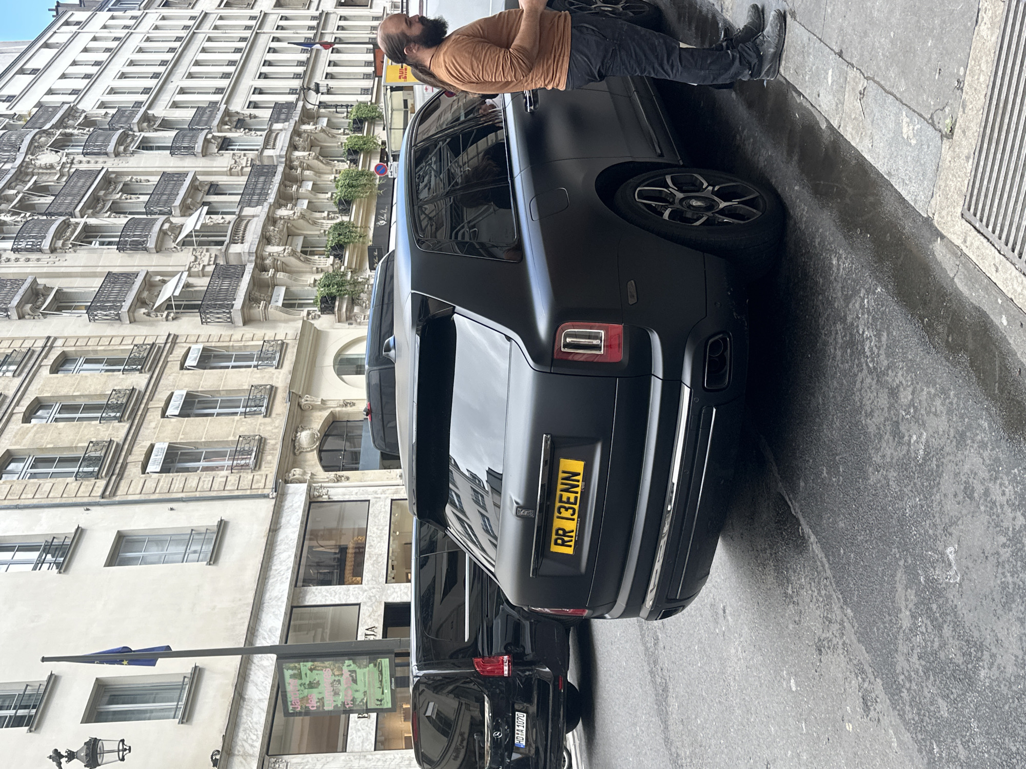 An eye-level outdoor shot in Paris, France, showing two black luxury vehicles parked on the street. A man with a beard and long hair stands next to the vehicle closest to the camera, partially obscured by the Rolls-Royce. The Rolls-Royce's license plate reads "RR 13ENN." The second vehicle, a black van, is partially visible and has a license plate of "HD-A1070." The architectural style of the surrounding buildings suggests a Parisian street with tall, light-colored buildings featuring windows, balconies, and ornate details. A street light pole and a sign with visible green and pink colors stand near the vehicles. The street surface shows signs of wear and is damp.