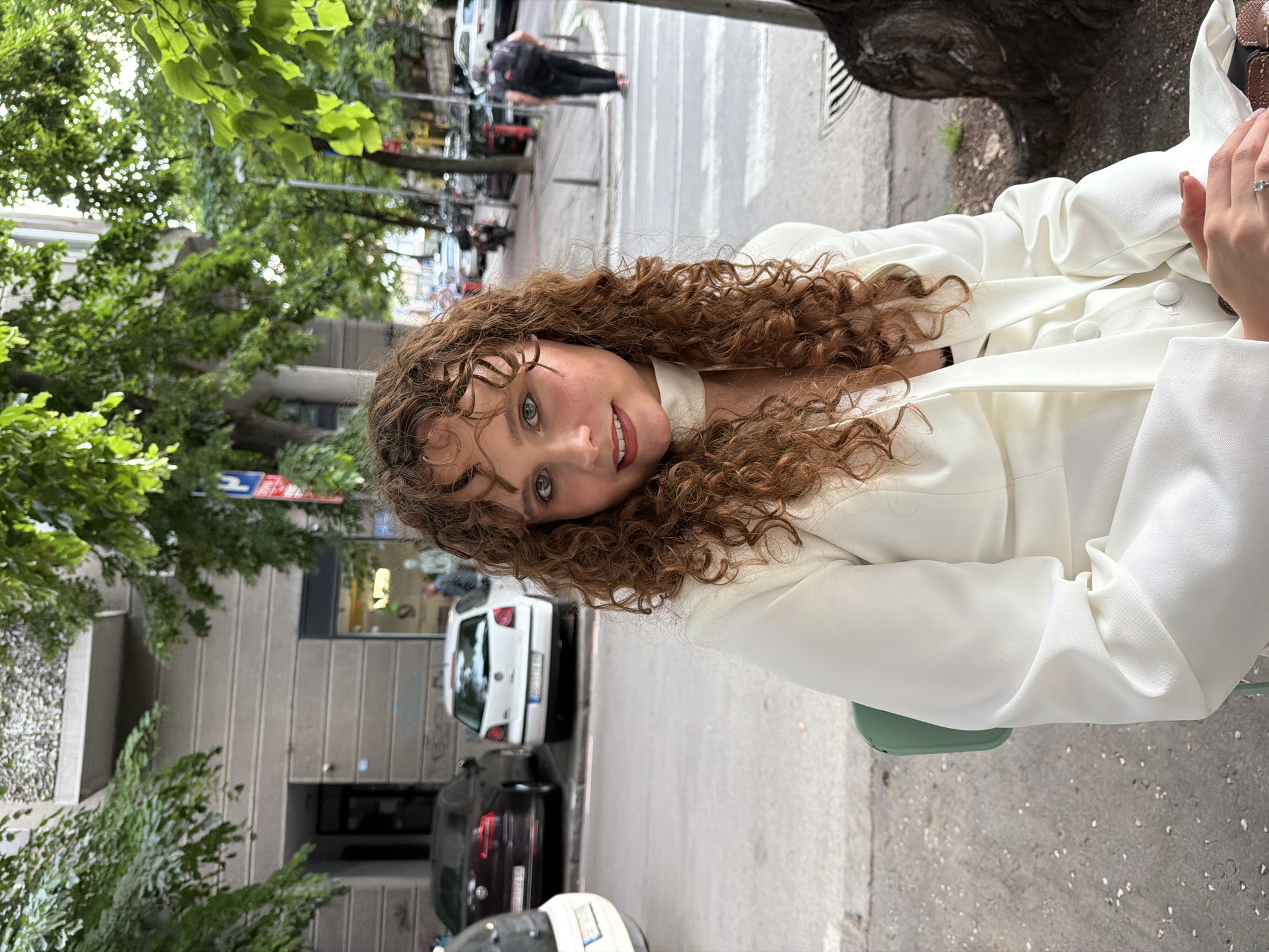 A woman with curly brown hair, wearing a white blazer, sits at an outdoor table in Belgrade, Serbia. The scene is an urban setting with trees lining the street and cars parked along the side. Pedestrians are walking in the background, and buildings can be seen across the street.