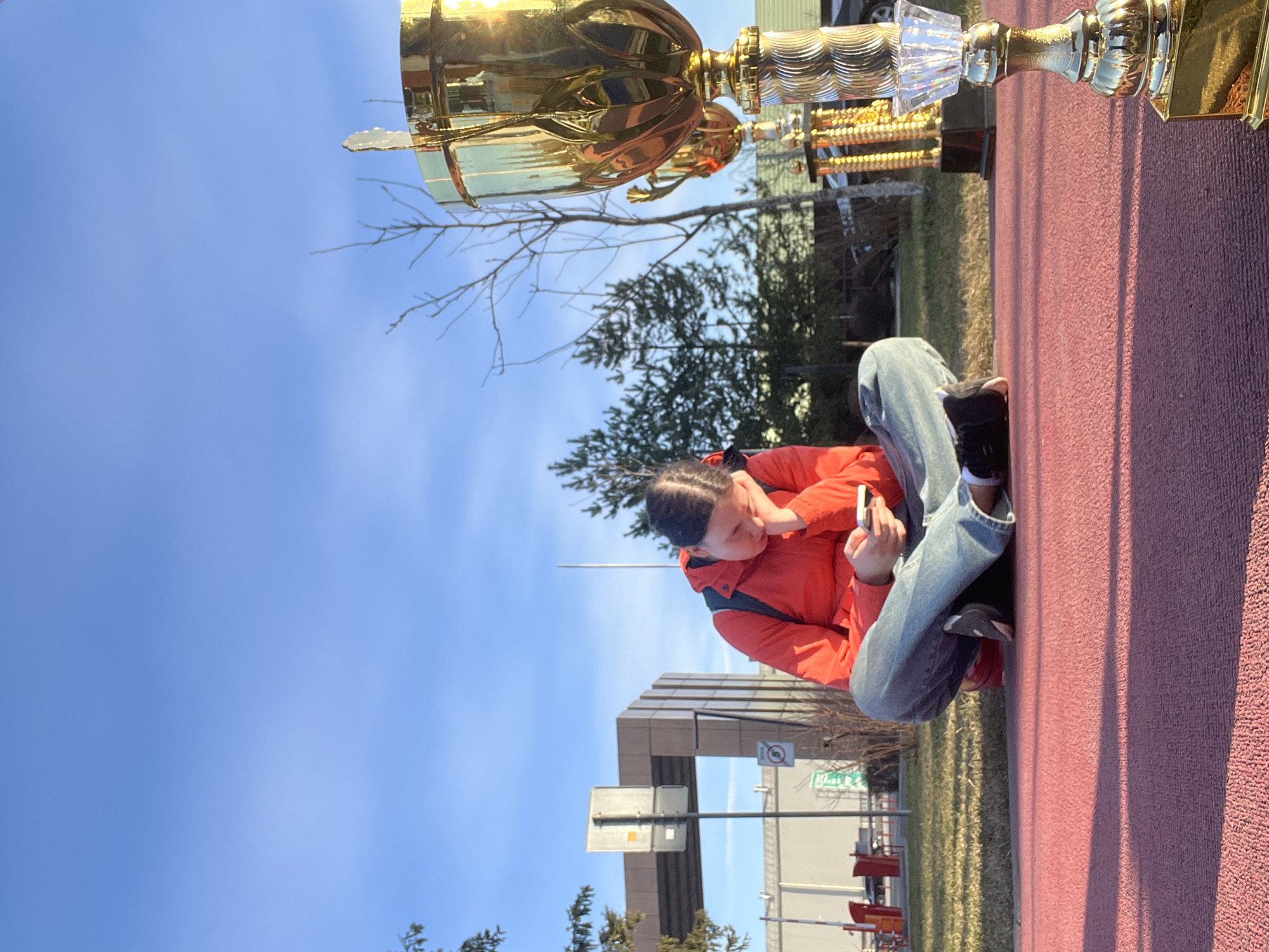 The image, oriented correctly, depicts a young person sitting on a red textured surface, possibly a running track, with a large golden trophy prominently displayed to their right. The person is dressed in a bright orange/red jacket, blue jeans, and sneakers, with their hair styled in braids. They are engrossed in their phone, which they hold in both hands.
The scene takes place outdoors on a sunny day under a clear blue sky. Bright sunlight casts distinct shadows, suggesting midday or early afternoon. In the background, bare tree branches indicate a late winter or early spring season. Further back, buildings with geometric architecture are visible, alongside other trees and hints of green grass. A blurred green street sign is partially visible in the middle ground, which, based on provided context, displays "Истра Гоу Доц". Another small, circular regulatory sign is also present, though its specific meaning is unclear. The trophy, highly reflective, captures sunlight and various elements of the surroundings, featuring a classic cup design with some clear, possibly crystalline, accents. The overall impression is one of a relaxed moment following an event or achievement, given the presence of the trophy.