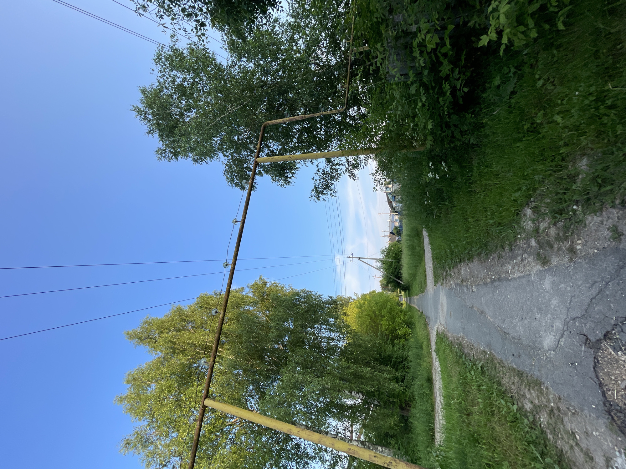 The image depicts a paved walkway in the village of Krasnye Baki, Russia, leading towards a treeline under a clear blue sky. Two yellow metal poles, part of a support structure for overhead wires, stand on either side of the walkway. The walkway appears worn, with cracks visible in the asphalt. Lush green grass and trees flank the path, with dense foliage on the right side of the frame. In the distance, power lines and utility poles are visible, along with a few houses with blue roofs, suggesting a residential area. The overall scene conveys a rural or semi-rural setting in the early evening or morning light. No people or animals are present in the image.
