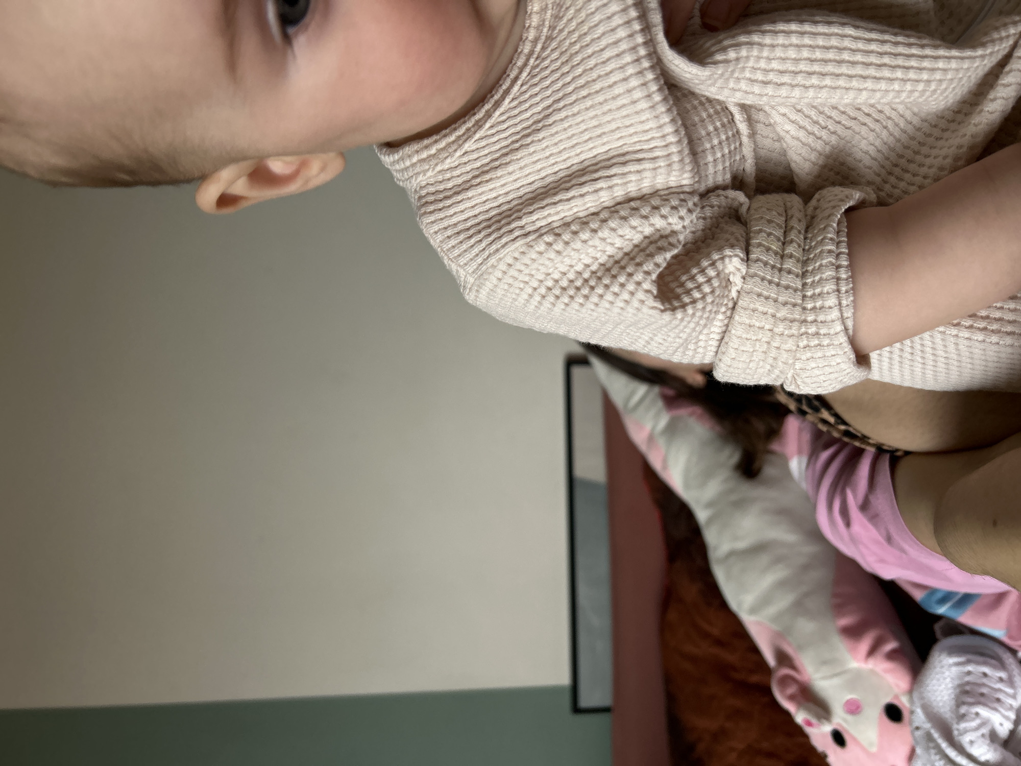 A close-up shot shows an infant wearing a light beige waffle-knit shirt. The infant's face, ear, and arm are visible. Beneath the infant is a multi-colored pillow, a pink shirt, and a white knitted item. The background consists of a two-tone wall, painted light grey above and muted green below, and a framed object. The scene is indoors. Location cannot be confirmed to be Moscow, Russia.