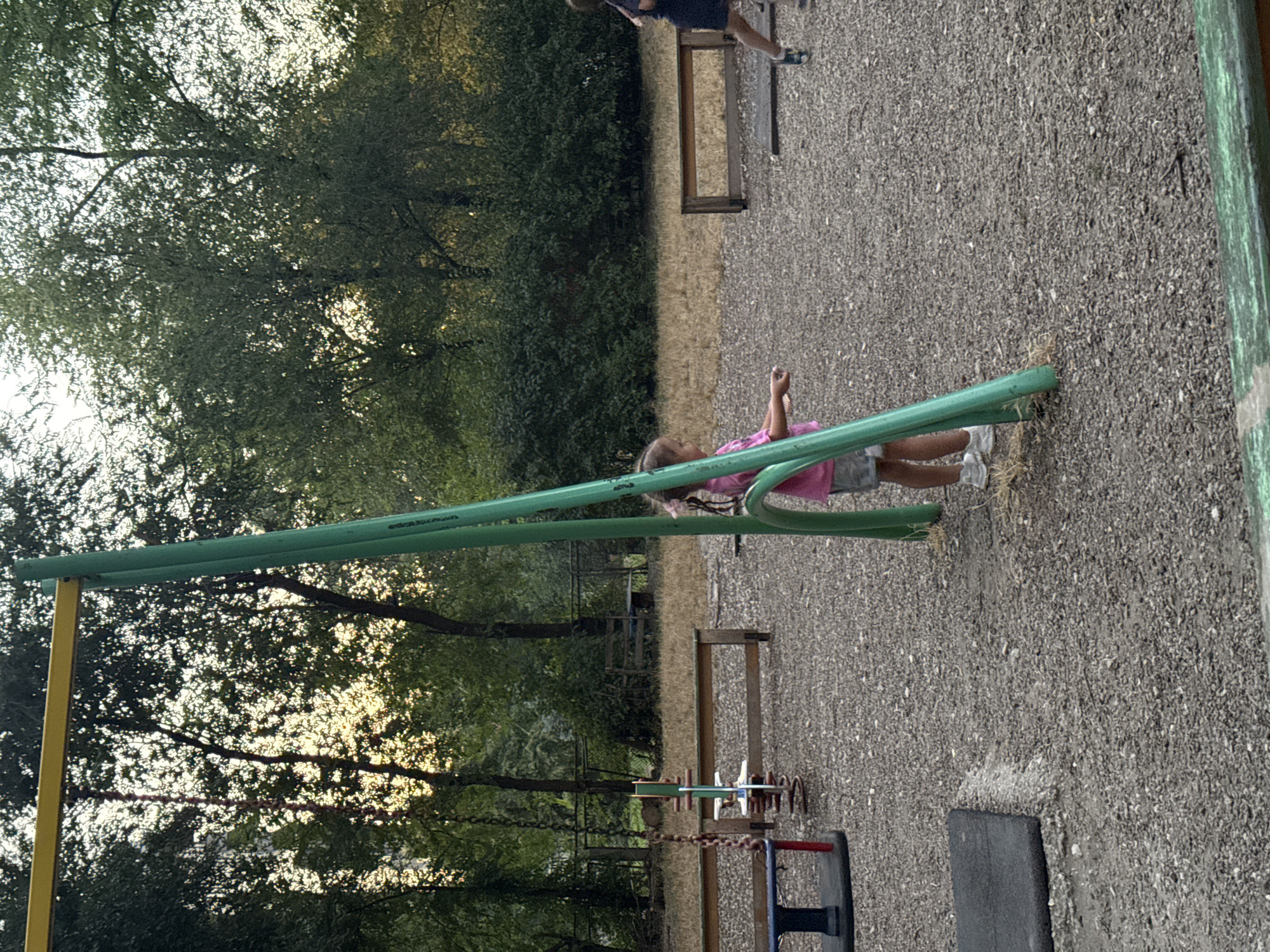This image depicts a playground scene in Chișinău, Moldova, during what appears to be late afternoon or early evening. In the foreground, a young girl with dark hair tied with a pink ribbon is standing near a tall, green metal support structure of a playground set. She is wearing a pink t-shirt and grey shorts, with white sneakers. Her right arm is extended, and she appears to be interacting with something out of frame. To the left, a swing set with a yellow crossbar is visible, with chains hanging down. Further into the playground, there are wooden benches and a ground covered in gravel and wood chips. In the background, dense green trees form a backdrop, with the sky visible through the foliage. A second, partially obscured figure, possibly another child, is visible on the right side of the image, in motion and wearing dark clothing. The overall impression is of a quiet, possibly residential, playground at the end of the day.