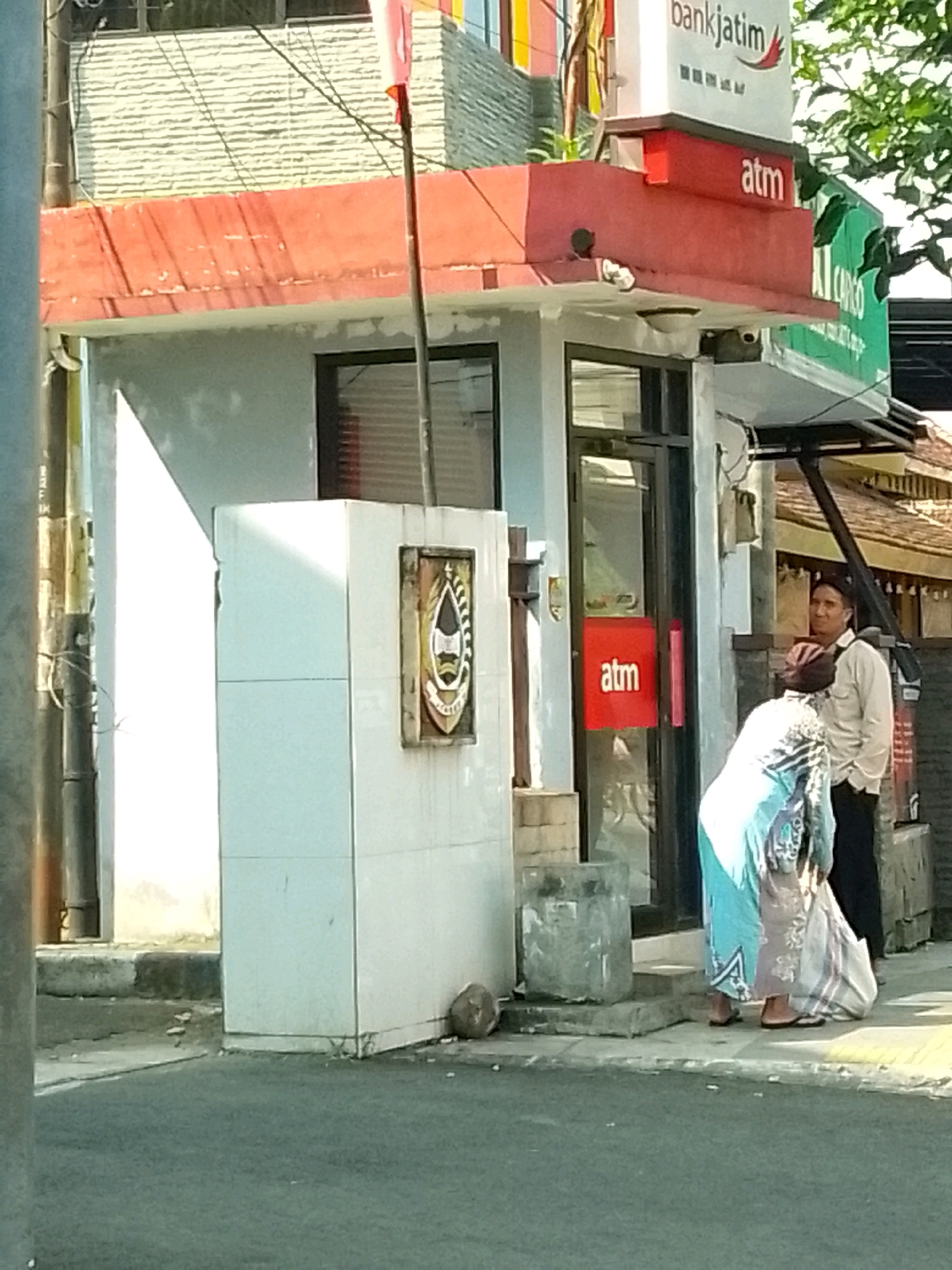 The image shows an outdoor scene in front of a bank building, possibly located in Jember, Indonesia. The bank building has a red awning with "atm" written on it in white lettering, and a prominent "bankjatim" logo at the top. To the left of the bank is a white tiled structure with an emblem displayed on it. Two individuals are visible in the foreground. One, appearing to be male, stands upright and looks to the right. The other, appearing to be female, is bent over, possibly adjusting or picking up items. Both individuals are dressed in casual attire. The environment suggests a sunny day with shadows cast on the ground. The scene includes a paved street, a sidewalk, and some vegetation in the background.
