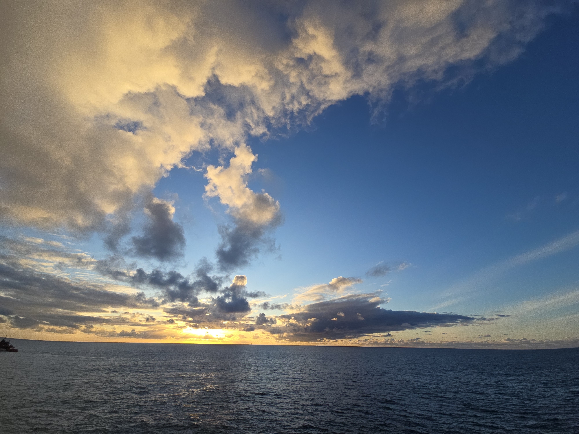The image depicts a seascape at sunset. The sky is a mix of blue and yellow-tinged clouds, with the sun low on the horizon. The sea is dark and calm, with gentle ripples on the surface. In the distance, a faint silhouette of a ship is visible on the left side of the frame. The location is an ocean scene of unknown origin, therefore I cannot determine the city.