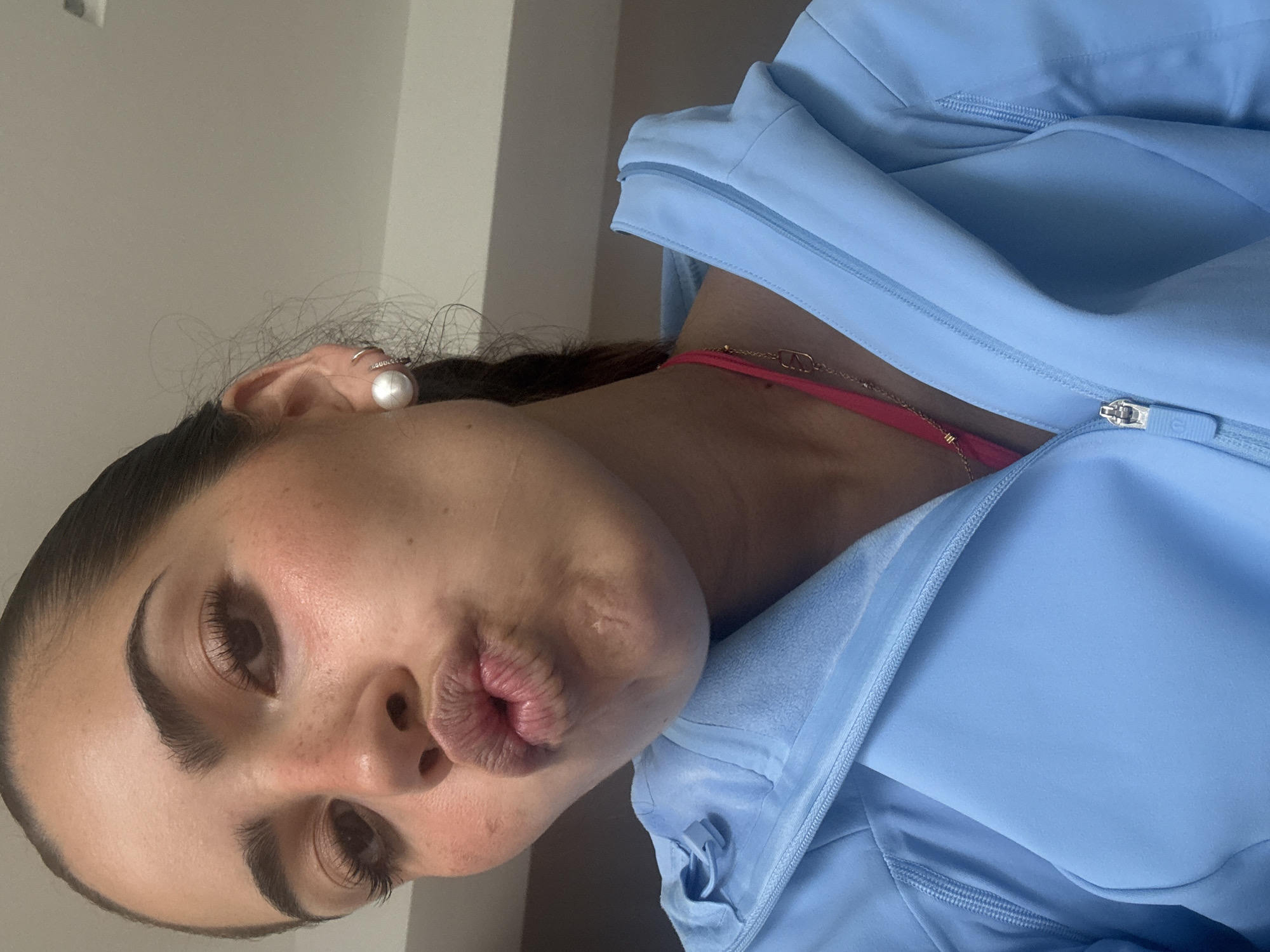 A young woman with dark hair pulled back is making a kissing face towards the camera. She is wearing a light blue zip-up jacket and a pink undergarment. She has on large pearl earrings and a delicate gold necklace. Her makeup is natural, with rosy cheeks and accentuated eyelashes. The background appears to be an indoor setting with neutral colored walls and ceiling. The image is a selfie, taken from a close-up perspective, focusing on her face and upper body. There is no information to suggest the location is San Bernardino Tlaxcalancingo, Mexico.