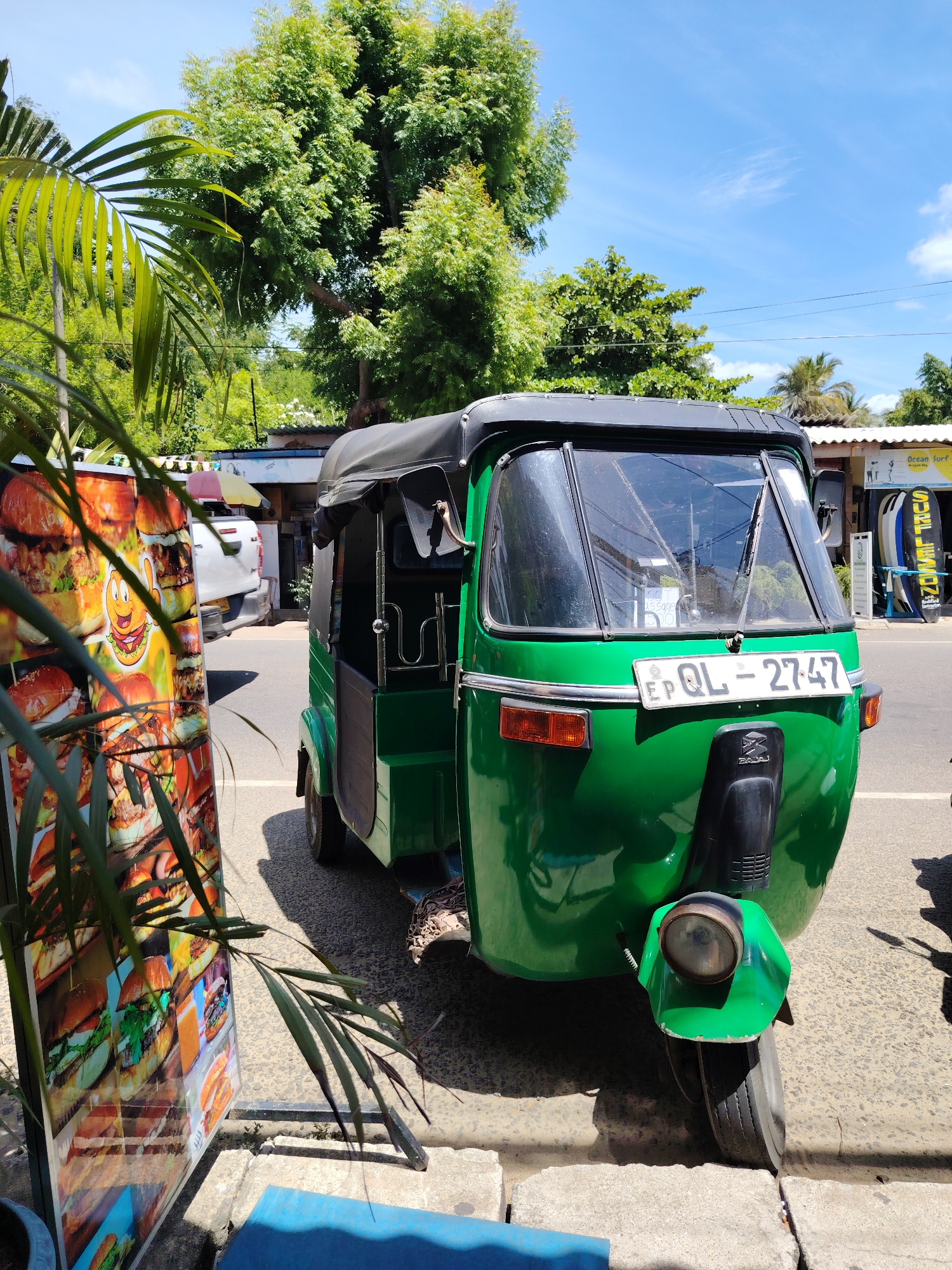 A green three-wheeled auto-rickshaw is parked on the side of a paved road. The auto-rickshaw has a black canvas roof and a license plate that reads "EP QL 2747". To the left of the auto-rickshaw is a large vertical advertising board featuring images of hamburgers. Behind the auto-rickshaw, a white pickup truck is visible. In the background, there are trees, a building with a white roof, and surfboards displayed outside a shop. The sky is blue with scattered white clouds. A palm frond is in the foreground, partially obscuring the view.