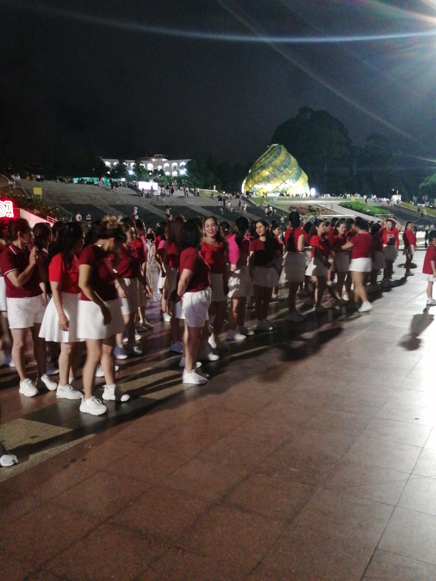 A group of women, likely performers, stand in a line on a paved surface at night in Da Lat, Vietnam. They are all wearing matching maroon polo shirts and white shorts or skirts, and white sneakers. Most of the women have dark hair, and some are smiling. In the background, a distinctive spiral-shaped building, illuminated with yellow and green lights, is visible. Beyond that, a large, dark hill is silhouetted against the night sky, with trees on its slopes. Further in the background, a large, white, multi-story building with arched windows is also illuminated, and a crowd of people can be seen gathered on tiered steps leading up to it. The overall atmosphere appears to be festive, possibly a performance or event taking place outdoors at night. The lighting suggests it is a clear night. There is no visible text in the image.