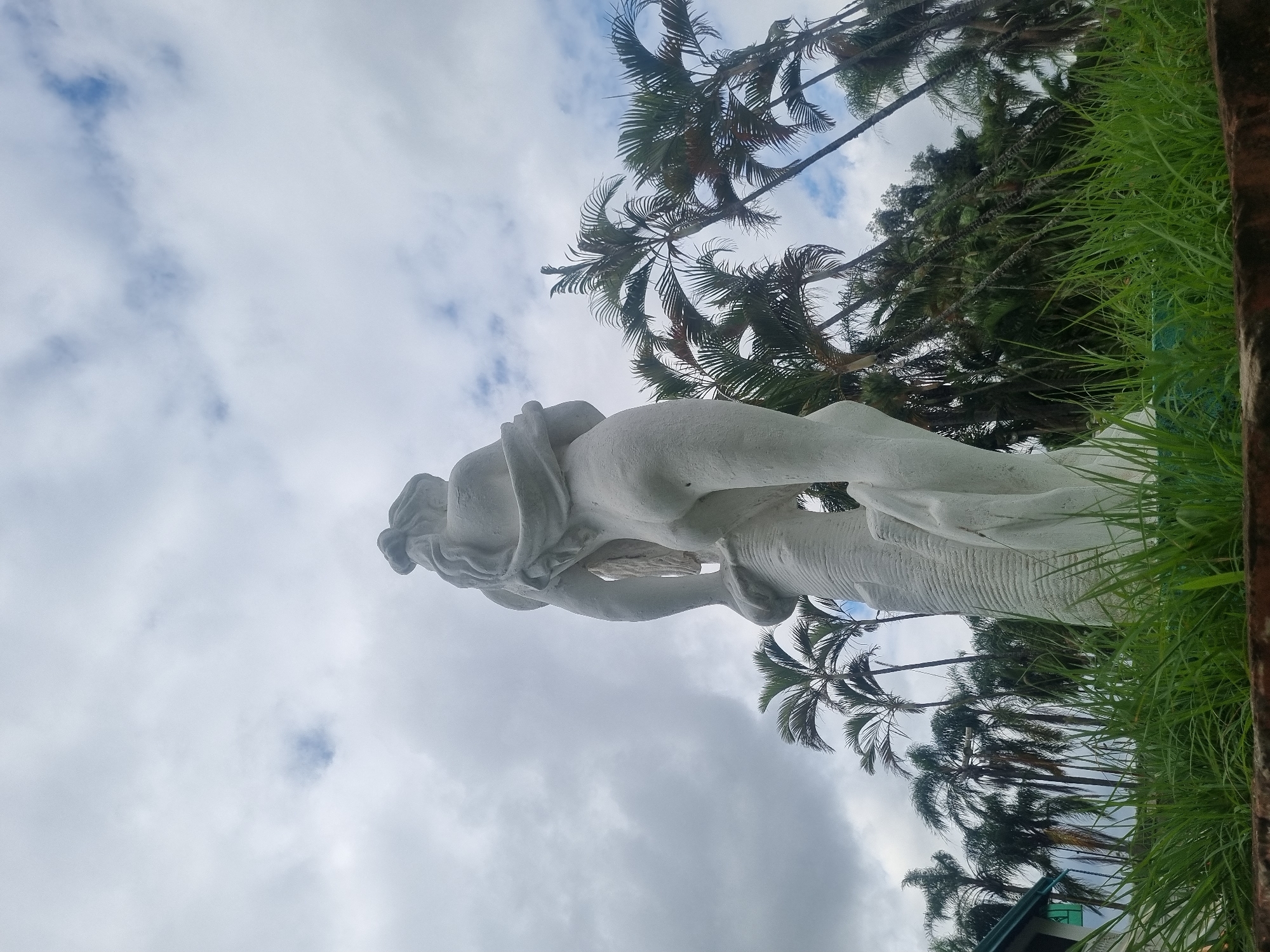 The image shows a white stone statue of a woman from behind, with her hair styled in braids. She is holding onto a textured column, and a piece of cloth is draped over her shoulder. The statue is set against a backdrop of a cloudy sky and numerous palm trees. In the foreground, lush green grass grows, and a section of a building with a green roof is visible to the left. The scene appears to be outdoors in a tropical or subtropical environment, possibly in the city of Laranjal Paulista, Brazil, given the presence of palm trees. The lighting suggests an overcast day.