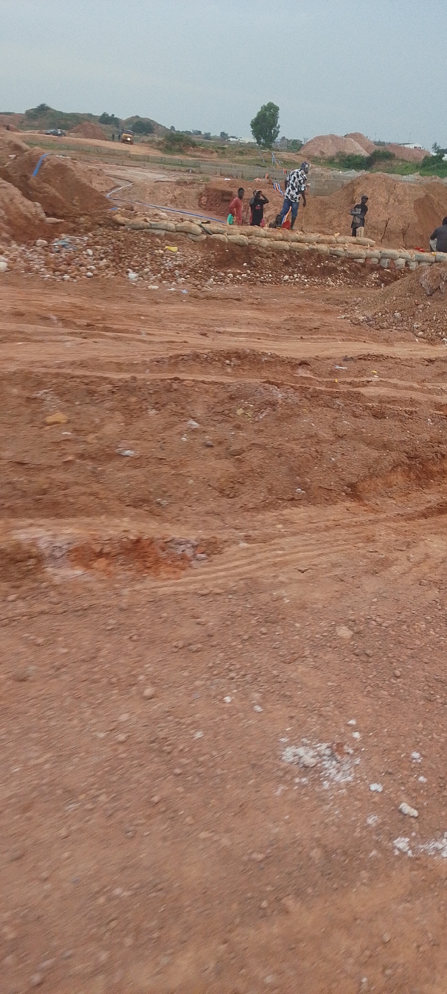 The image shows a construction site, likely in Rayfield, Nigeria, with several individuals present. The foreground is dominated by reddish-brown soil, indicative of earthworks. In the middle ground, sandbags are arranged to form a barrier, behind which several people are visible. One individual, wearing a patterned shirt and blue jeans, stands facing away from the camera. To their left, two other figures appear to be in conversation, one in a red shirt and the other in a dark shirt. Further to the right, another person in a dark shirt stands. The background reveals further construction activities, including mounds of soil, a truck, and some vegetation. The sky is overcast.