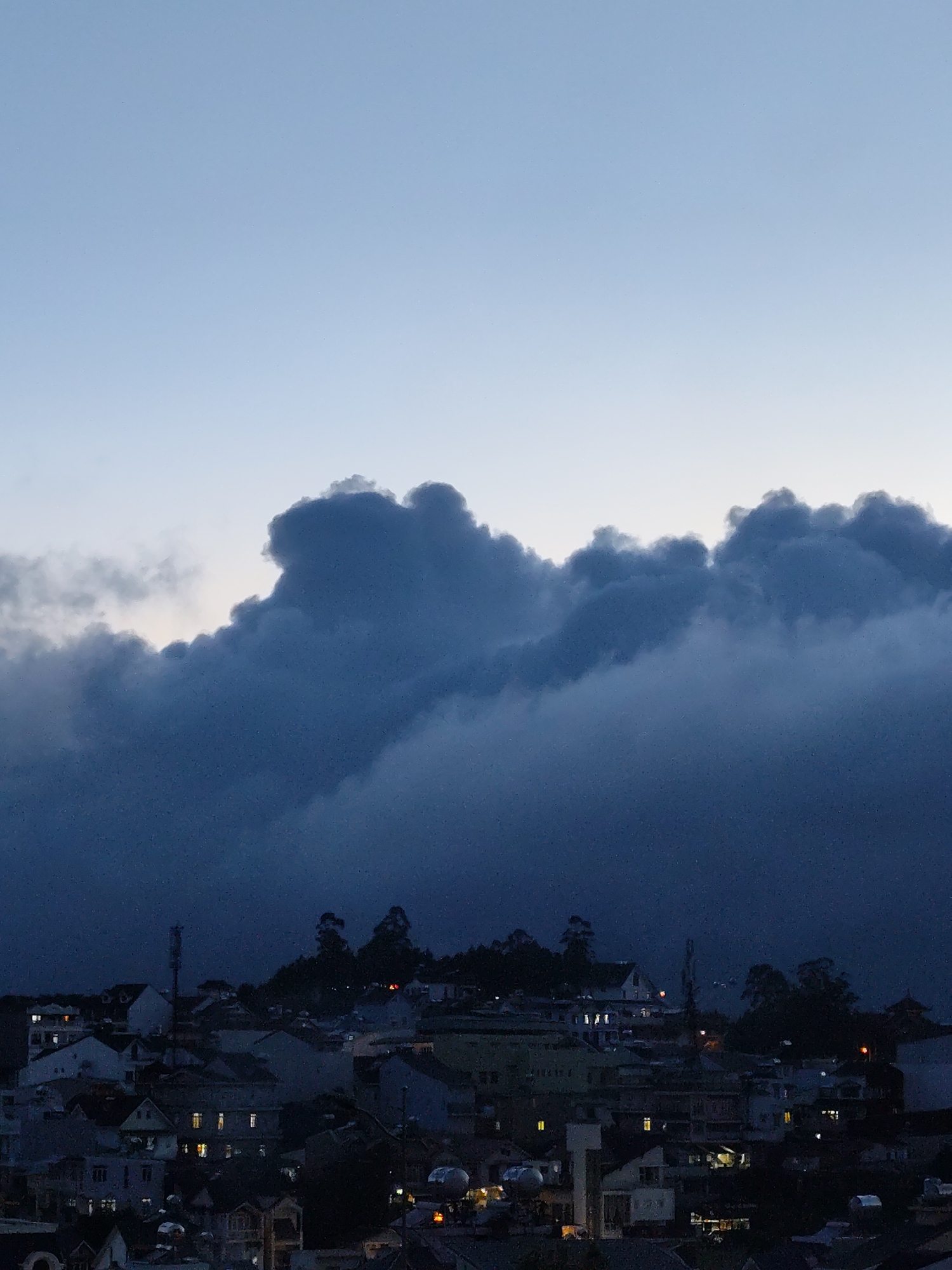 The image shows a view of Lang Biang - Da Lat, Vietnam, at dusk. In the foreground, there is a dense collection of buildings, with the roofs and facades dimly lit by scattered lights. Behind the buildings, a row of trees and a communication tower can be seen on a higher ground. The background is dominated by a large, dark cloud formation that fills the upper half of the frame. The sky above the clouds fades from light blue to a darker shade.