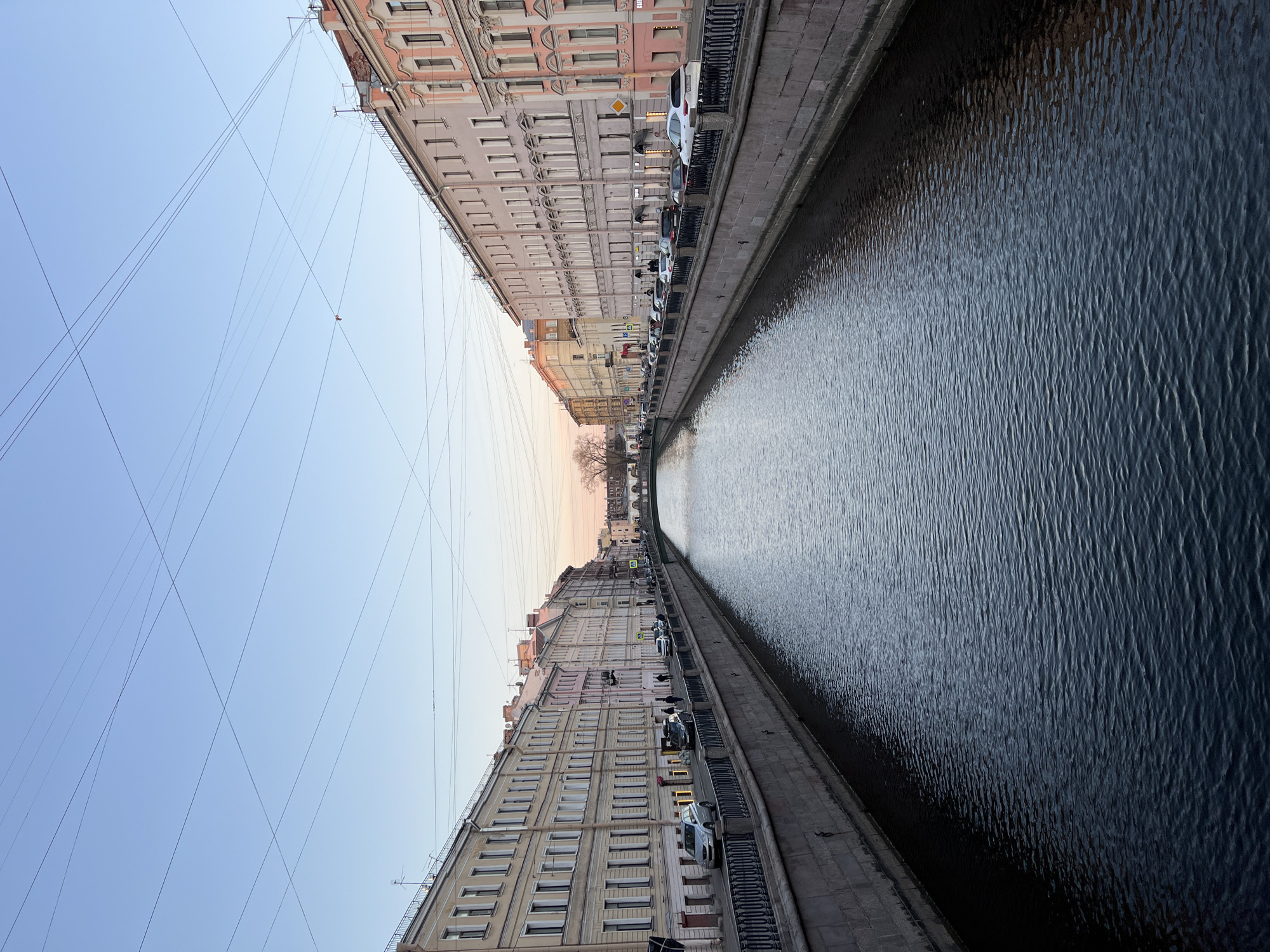 This image depicts a canal scene in Saint Petersburg, Russia. Tall buildings line both sides of the canal, with parked cars along the canal edge. Overhead power lines crisscross above the buildings. The water in the canal is dark with visible surface ripples. The sky is a light blue color, suggesting either early morning or late afternoon.