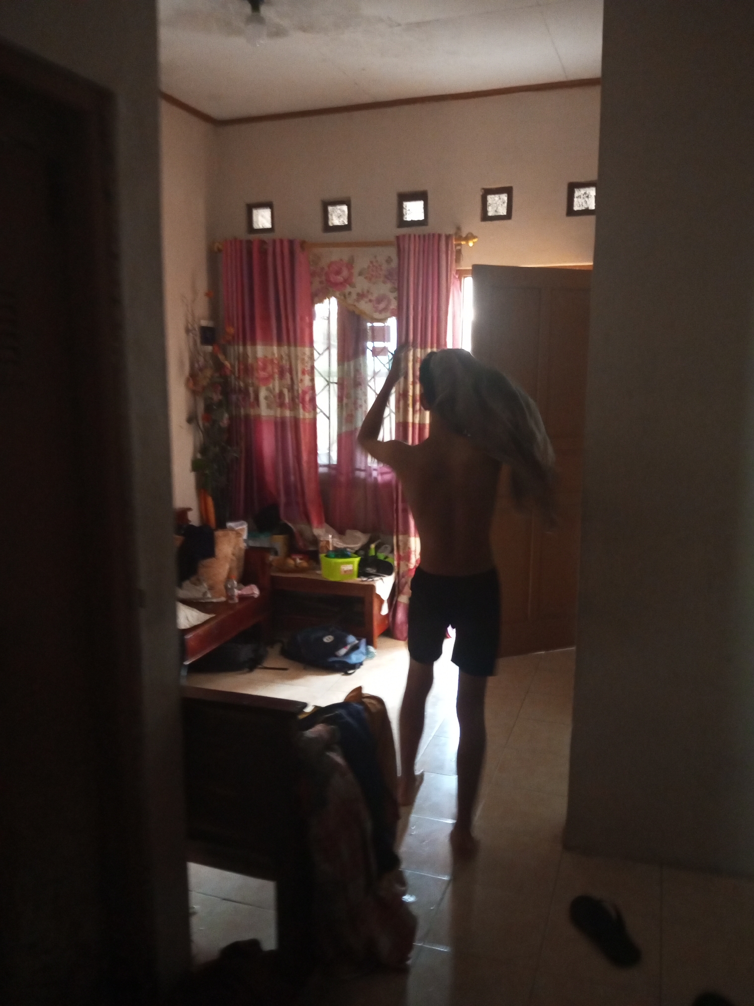 The image depicts an indoor scene within a room in Banyudono, Indonesia. The primary subject is a young man, seen from behind, who appears to be drying his hair with a towel. He is shirtless and wearing dark shorts. The room is somewhat cluttered, with items visible on shelves, furniture, and the floor. Red curtains with floral patterns hang across a window, and there are decorative square elements on the wall above the curtains. The lighting suggests it could be daytime, with natural light coming through the window. The overall impression is of a casual, lived-in space, possibly a bedroom or living area. The presence of a ceiling fan, the style of the curtains, and the tiled floor are common features found in Indonesian homes. There is no visible text in the image.