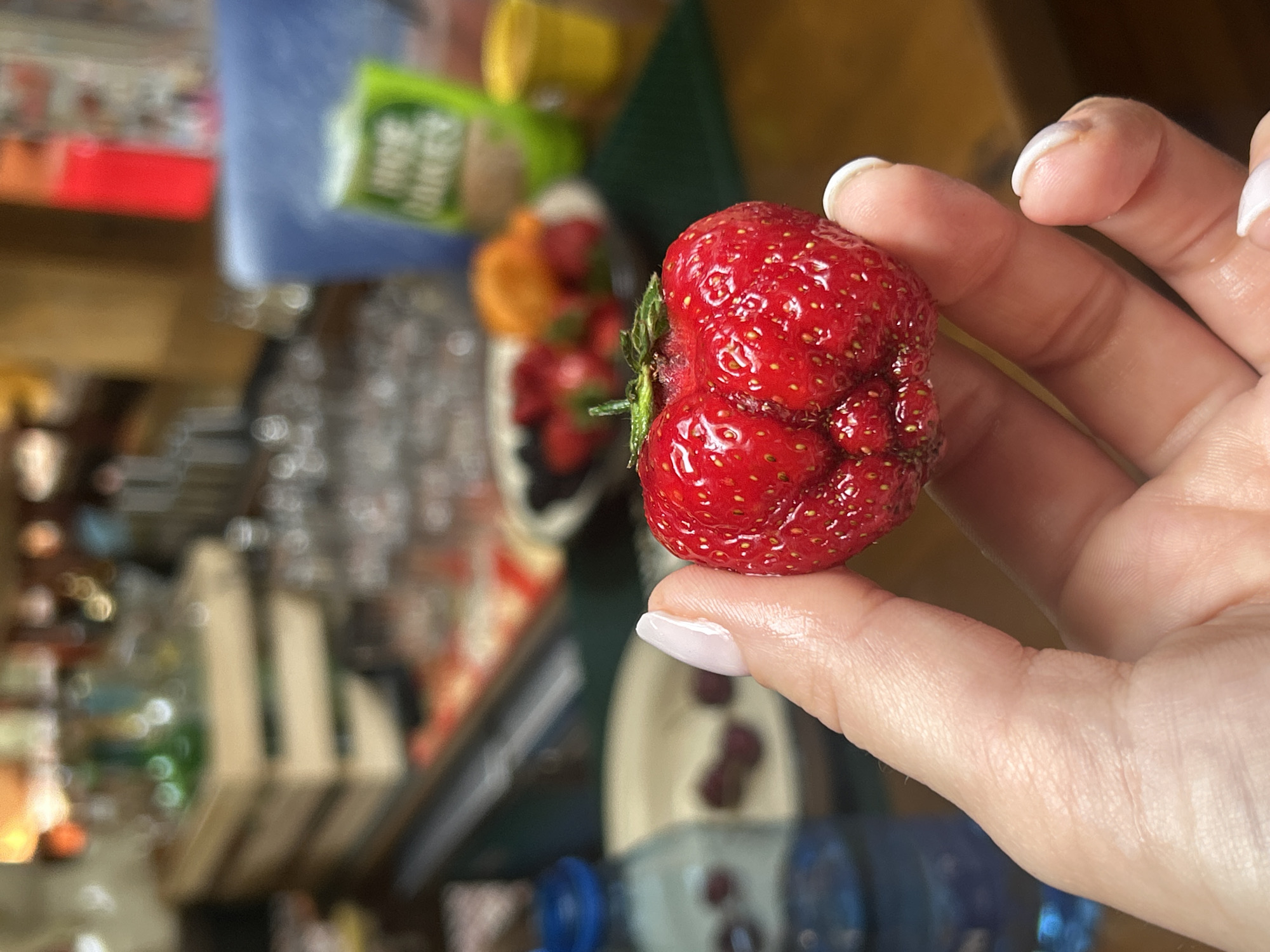 In the foreground, a person's hand, with well-maintained light-colored fingernails, holds a large, misshapen strawberry. The strawberry is bright red and has visible seeds. In the background, the scene is blurred but suggests an indoor setting with various objects. There are hints of what might be packaging with green and white text, possibly food-related. Other items appear to be containers and possibly more fruit. The overall impression is that of a casual moment, perhaps during a meal or snack. The lighting suggests daytime.