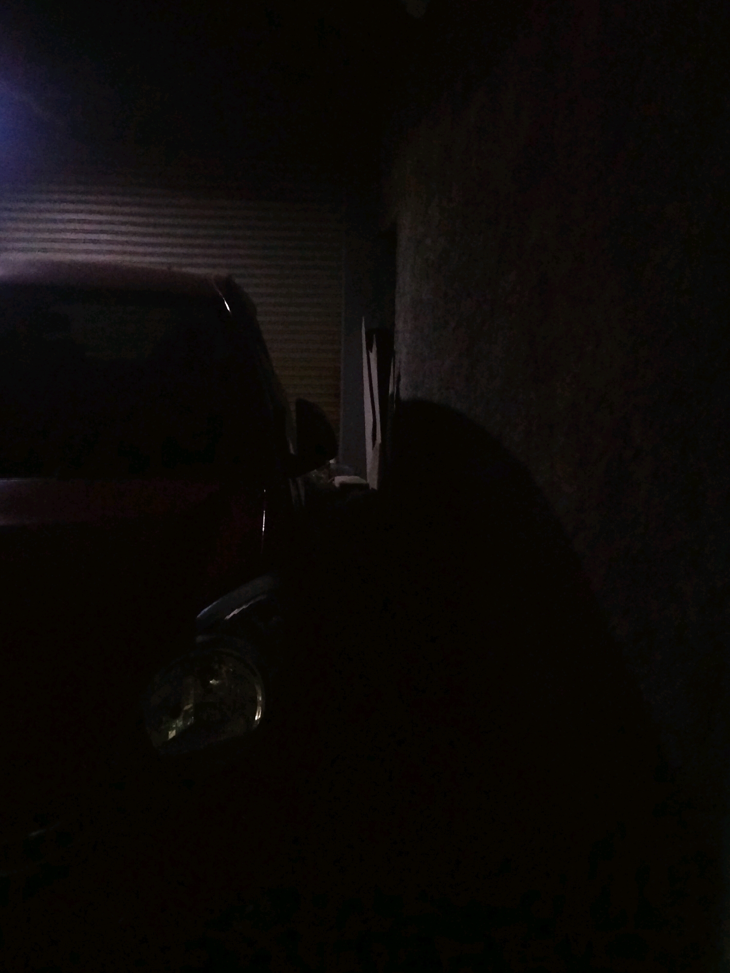 The image depicts a dimly lit scene, likely at night, within a garage or covered area. On the left, a portion of a dark-colored vehicle is visible, with its headlight illuminated. The headlight's lens reflects light, suggesting it is on. Behind the vehicle, a corrugated metal surface is apparent, indicating a garage door or wall. To the right of the vehicle, a dark, textured wall rises, casting a shadow that extends across the scene. The ground is not clearly visible due to the low light conditions. The overall impression is one of a parked vehicle in an enclosed space, possibly a residence in Suka Sari, Indonesia.