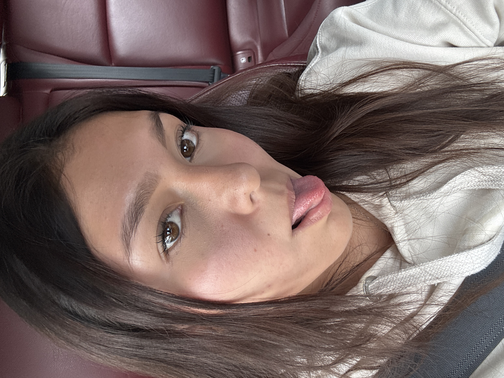 A young woman with brown hair is sticking her tongue out. She is wearing a white hoodie. She is sitting in a car with red leather seats and a seatbelt. The exact location in Oaxaca City, Mexico cannot be determined from this image.