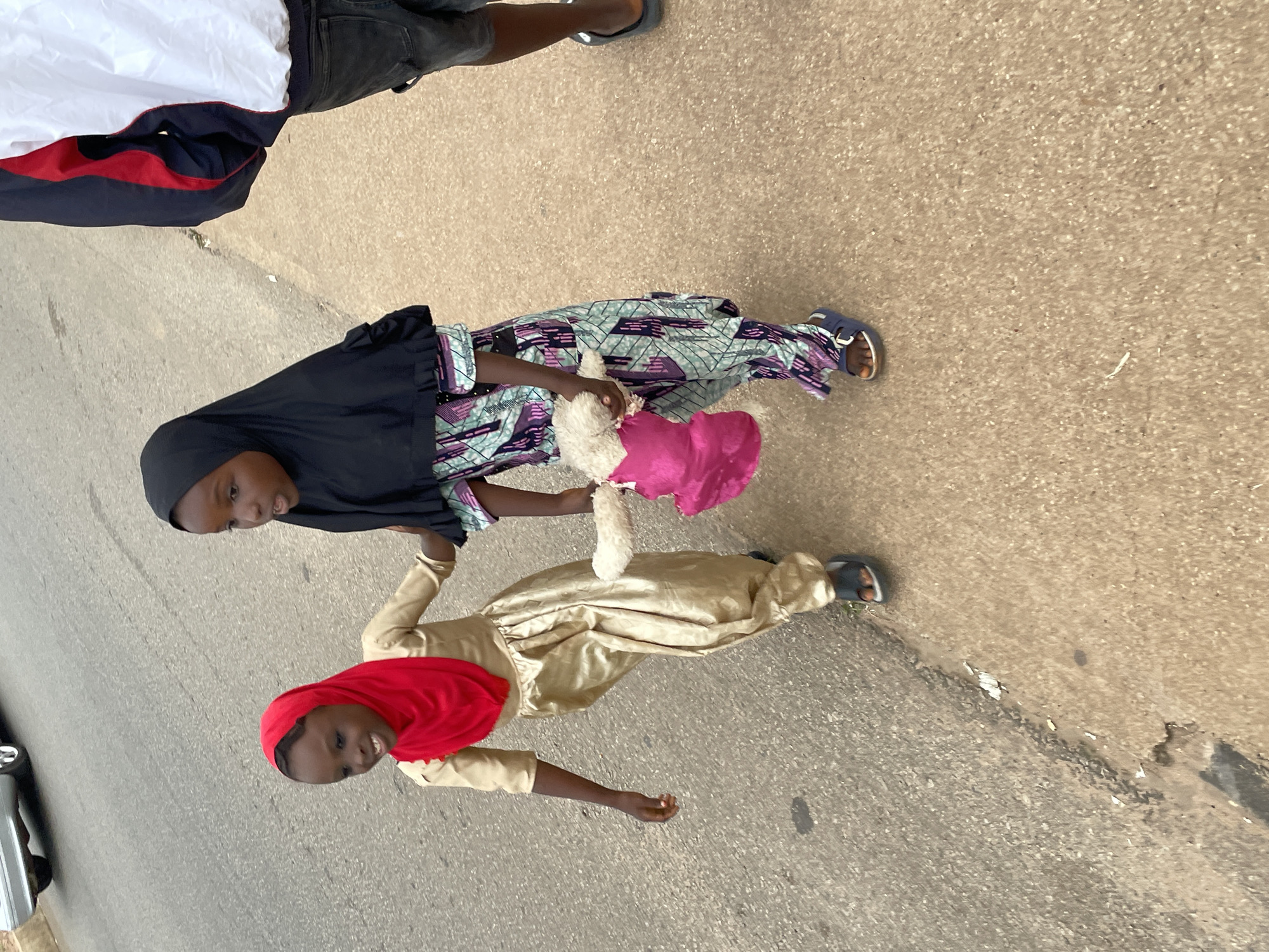The image shows two young girls standing on a paved surface, likely a street or sidewalk in Jos, Nigeria. The girl in the foreground is wearing a gold dress and a red hijab, smiling at the camera. She is holding a plush toy. Behind her, another girl is wearing a black hijab and a patterned dress. She is also looking towards the camera, holding the plush toy with the other girl. The ground appears to be asphalt with some debris. The lighting suggests it is daytime, possibly with overcast or diffused sunlight. The overall mood appears cheerful, with both girls smiling.