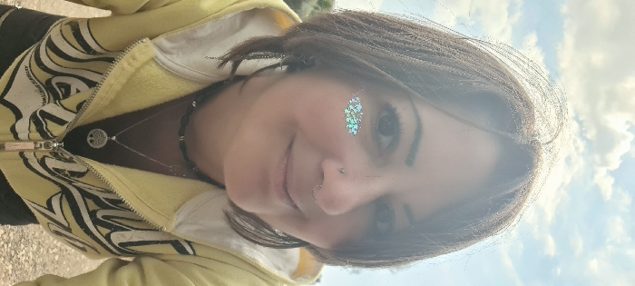 The image displays an adult female depicted in a close-up, slightly upward-angled outdoor portrait. She has medium-length brown hair and is looking directly at the camera with a subtle, closed-mouth smile. Her left eye (viewer's left) has a patch of holographic glitter applied beneath it. She wears a silver stud piercing in her left nostril and a silver ring piercing in her septum. Visible accessories include a chain-style earring on her left ear, a black beaded necklace, and a longer silver-toned necklace with a circular "Tree of Life" pendant. She is dressed in a yellow zippered hoodie featuring black and white graphic elements, worn over a black shirt. The background consists of a blue sky with scattered white clouds, with faint green foliage discernible on the left side. The ground, visible at the bottom of the frame, appears light-colored. The location is the Mate Asher Regional Council, Israel.