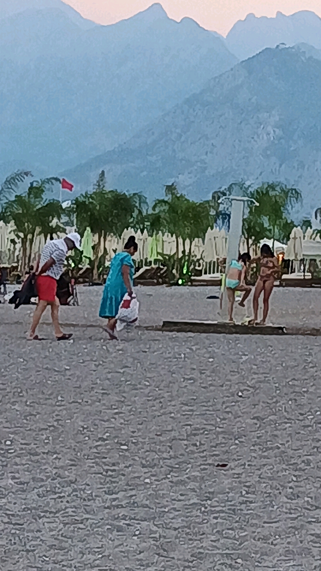 In Konyaaltı, Turkey, a beach scene unfolds under a hazy, mountainous backdrop. A man in a striped shirt and red shorts walks on the pebbled shore, carrying a dark bag. Beside him, a woman in a blue dress walks with a large white bag. Further along the beach, two children, one in a light blue bikini and the other in a darker bikini, are near a public shower. Behind them, beach chairs and umbrellas are arranged. The overall atmosphere is one of casual beach activity.