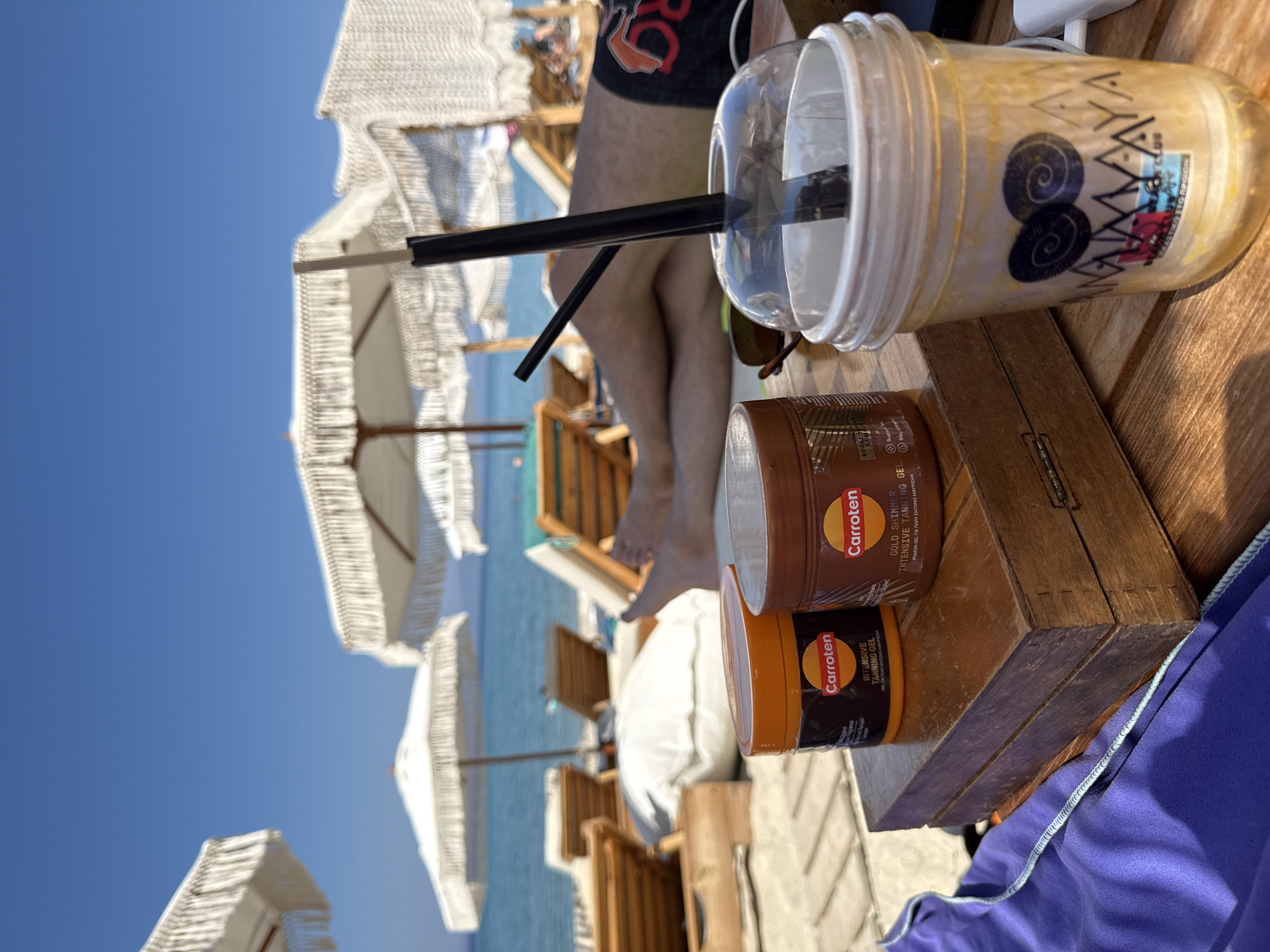 This image depicts a sunny day at the beach, likely in Greece given the location context. The scene is set on a wooden table outdoors, with white beach umbrellas and the blue sea visible in the background.
In the foreground, there are several objects: a clear plastic cup with a beverage and a black straw, two orange and brown jars of Carroten tanning gel, and a pair of sunglasses. The visible text on the jars includes "Carroten", "INTENSIVE TANNING GEL", and "GOLD SHIMMER". One jar also has Greek text indicating "GEL ΓΙΑ ΠΟΛΥ ΕΝΤΟΝΟ ΜΑΥΡΙΣΜΑ" which translates to "GEL FOR VERY INTENSE TANNING".
A person's legs and torso are visible, suggesting they are relaxing on a beach chair, possibly enjoying the drink and preparing to tan. The bright sunlight and clear sky indicate it is daytime, and the setting suggests a vacation or leisure activity. The overall mood is relaxed and summery.