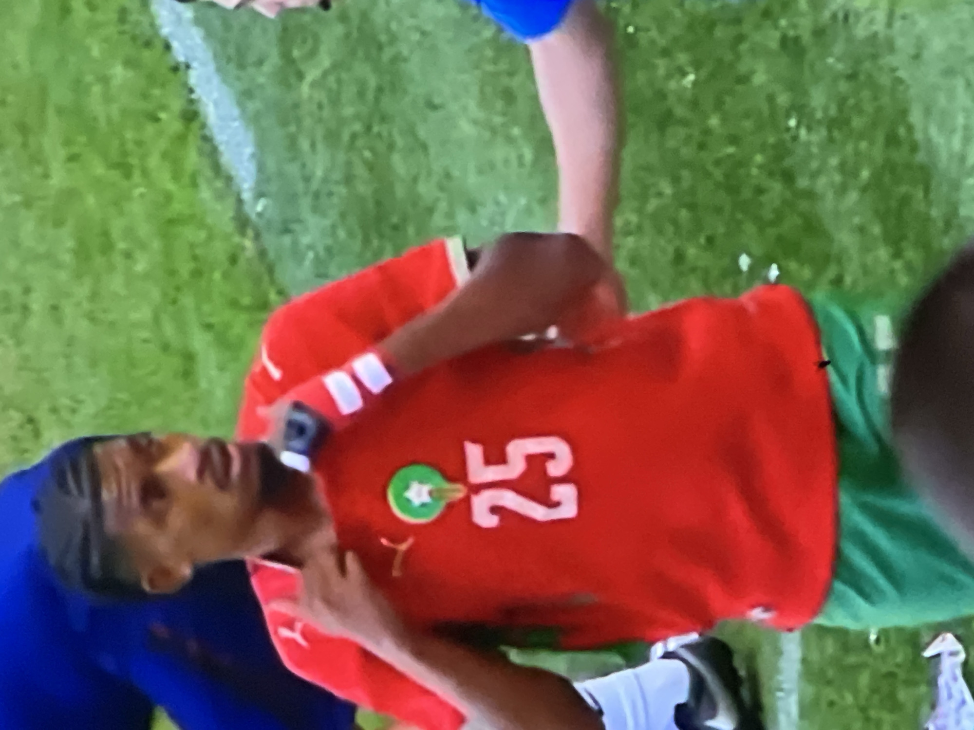 A male individual, identified as a football (soccer) player wearing a red short-sleeved jersey and green shorts, is depicted on a green grass field marked with a white line. The player has dark skin, a trimmed beard and mustache, and dark hair styled in short braids or cornrows on top with faded sides. He is looking towards his right (viewer's left) with a slight open-mouthed expression.
The red jersey features a yellow Puma logo on the right chest. On the left chest, the emblem of the Royal Moroccan Football Federation (FRMF) is visible, consisting of a green circle with a white star and a crown-like symbol above it. Below this emblem, the number "25" is prominently displayed in white. The player's left hand is near his collar, while his right hand holds a small black object, possibly a smart device, with white athletic tape wrapped around his wrist.
To the right of the main subject, a Caucasian forearm wearing a blue sleeve is partially visible, extending towards the player, suggesting an interaction such as a handshake or touch. In the background, to the left, parts of another individual in a dark blue garment and white socks with black shoes (a Nike swoosh is faintly visible) are present.
The environment is an outdoor football pitch, characterized by green grass and a visible white sideline. The lighting suggests daylight conditions. While the player's attire indicates representation of Morocco, the specific location in Casablanca, Morocco, cannot be confirmed solely from the visual information within this image.