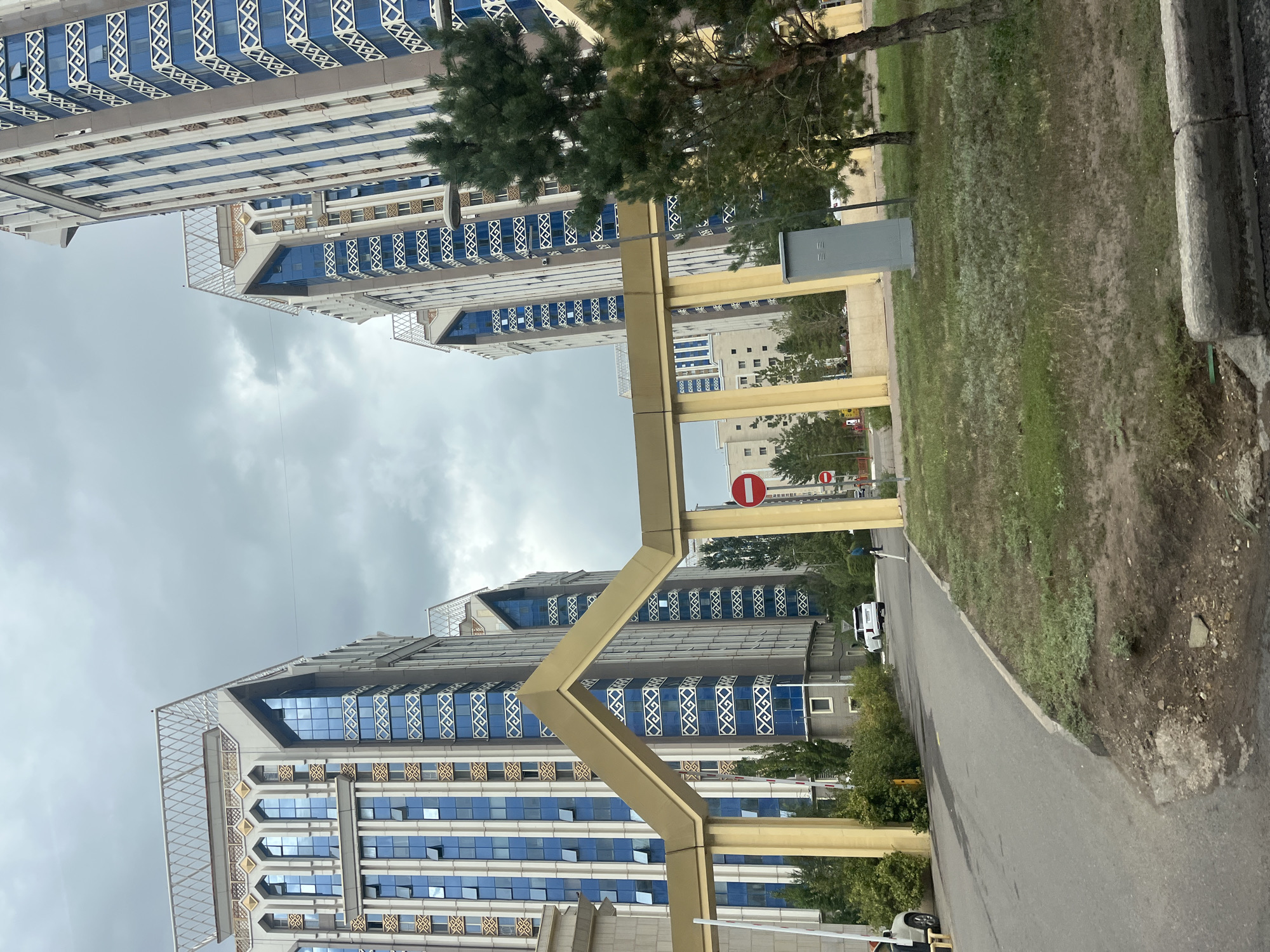 The image depicts a street scene in Astana, Kazakhstan, characterized by modern, high-rise apartment buildings with blue glass facades and ornate architectural details. A golden archway structure frames the view of the buildings and a road leading into a complex. The sky is overcast, suggesting a cloudy day. On the road, a white SUV is parked, and a person is walking away from the camera. Green trees and grass are visible in the foreground and along the sides of the road. Traffic signs, including a red circular sign with a horizontal white bar, are present. A grey utility box is situated on the grassy area to the right of the road. The architecture suggests a contemporary residential area within the city.