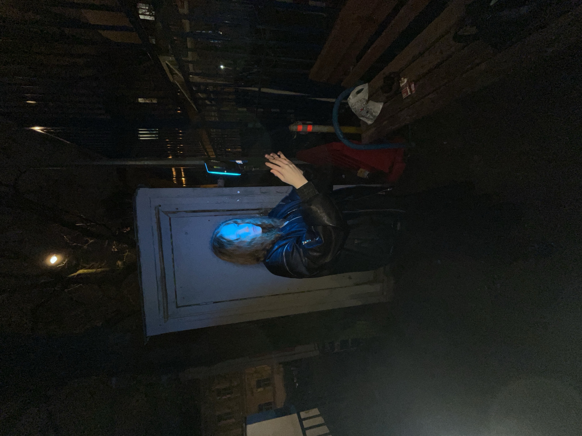 A person with long, wavy blonde hair lies on a white surface, possibly a door or large panel, illuminated by a bright blue light on their face. The person is wearing a black leather jacket. The surroundings are dark, suggesting a nighttime setting. To the right, there are some wooden planks and various objects, including what appears to be a red container, a white object with a design, and a blue hose. In the background, there's a dim streetlight on the left and a building partially visible at the bottom. The location is indiscernible from the image.