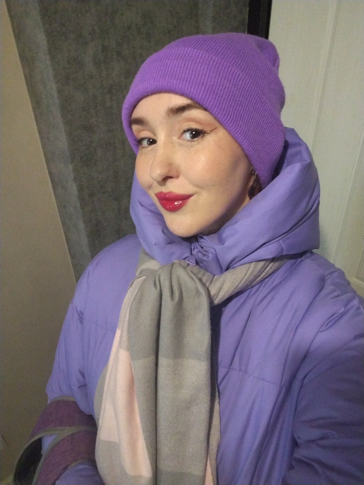 A person is shown in a selfie style wearing a lavender puffer jacket, a matching purple beanie, and a gray and pink scarf. The background appears to be indoors with a gray wall and a white paneled door visible. There's no information available in the image to connect it to Izhevsk, Russia.