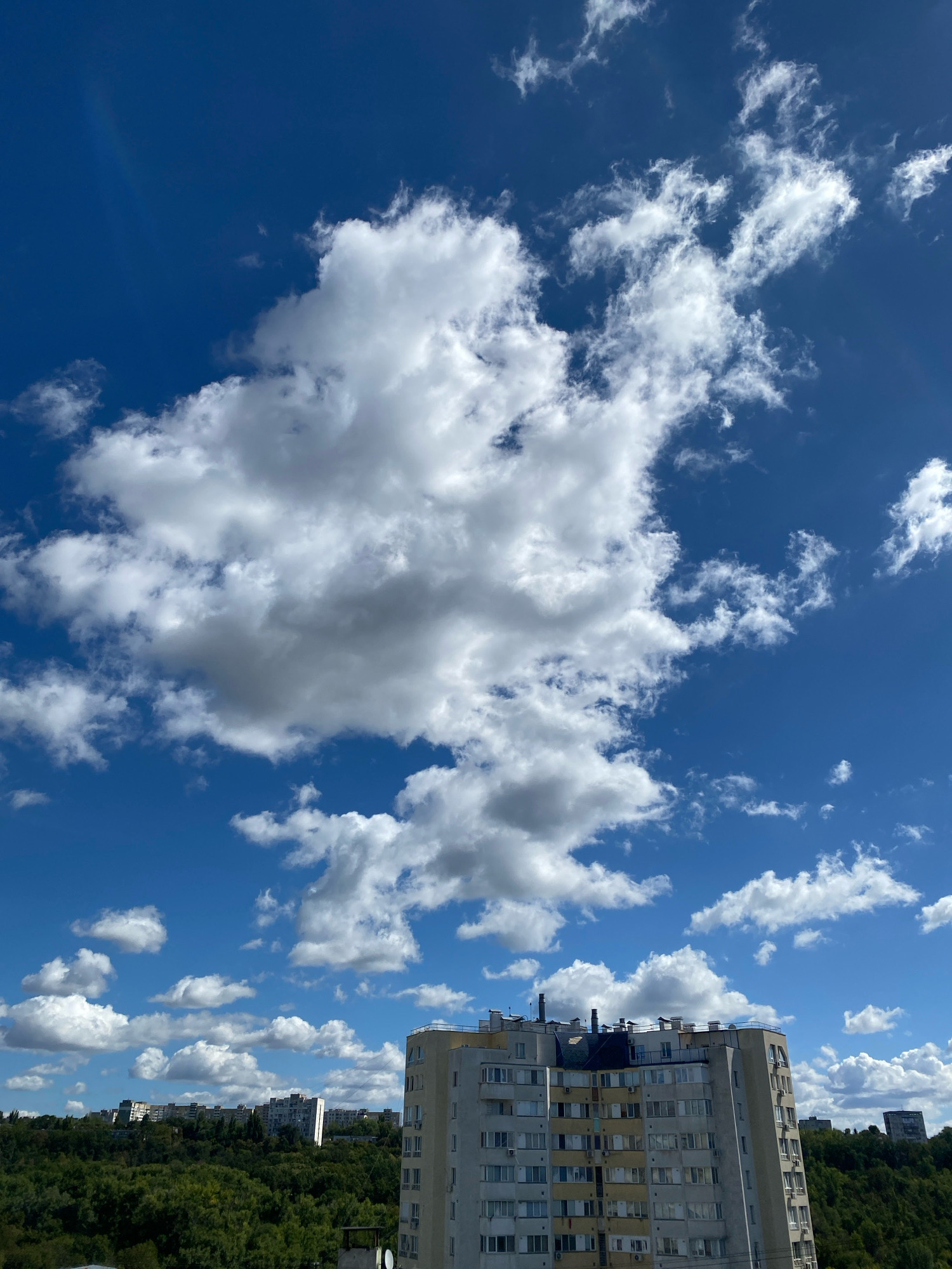The image captures a sky view over a residential area in Chișinău, Moldova, on a bright, partly cloudy day. The upper portion of the frame is dominated by a vivid blue sky with scattered cumulus clouds of varying sizes and densities. One large, prominent cloud formation sits towards the center-left, with other smaller clouds dispersed throughout the sky. In the lower portion of the image, a multi-story apartment building with a light-colored facade and numerous windows is visible on the right. Behind the building and extending across the middle ground, a dense canopy of green trees covers a hillside. In the distance, several other apartment buildings are visible among the trees, suggesting a developed urban landscape. The overall scene depicts a typical urban environment under a dynamic sky.