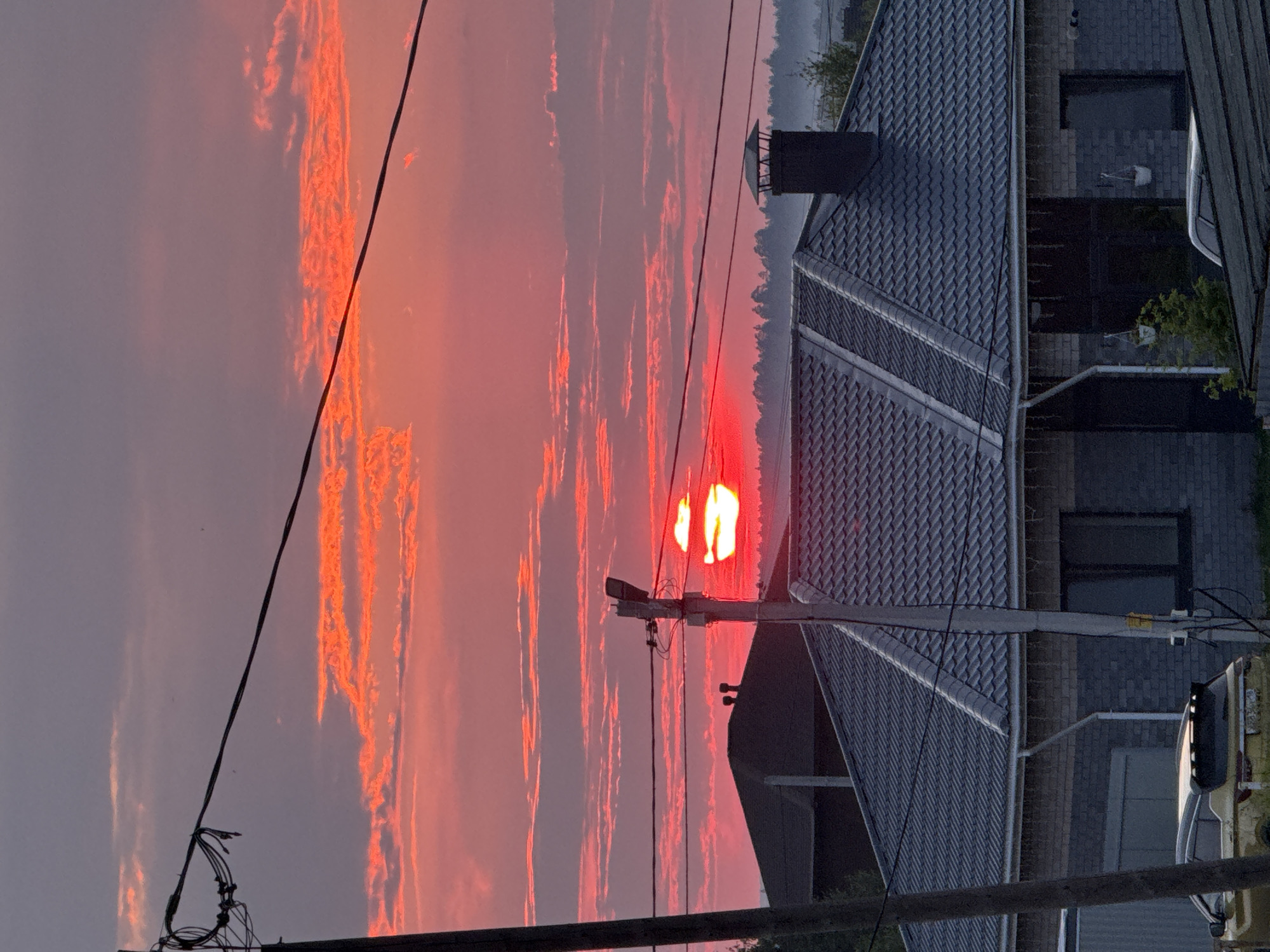 The image captures a vivid sunset over a suburban area in Izhevsk, Russia. The sky is a dramatic canvas of pink and orange hues, with clouds illuminated by the setting sun, which appears as a bright red orb partially obscured by a utility pole. In the foreground, a modern house with a grey tiled roof and brick exterior is visible, featuring a prominent chimney. A yellow car is parked in front of the house. Power lines stretch across the frame, adding to the urban landscape. The overall scene suggests a tranquil evening in a residential district.