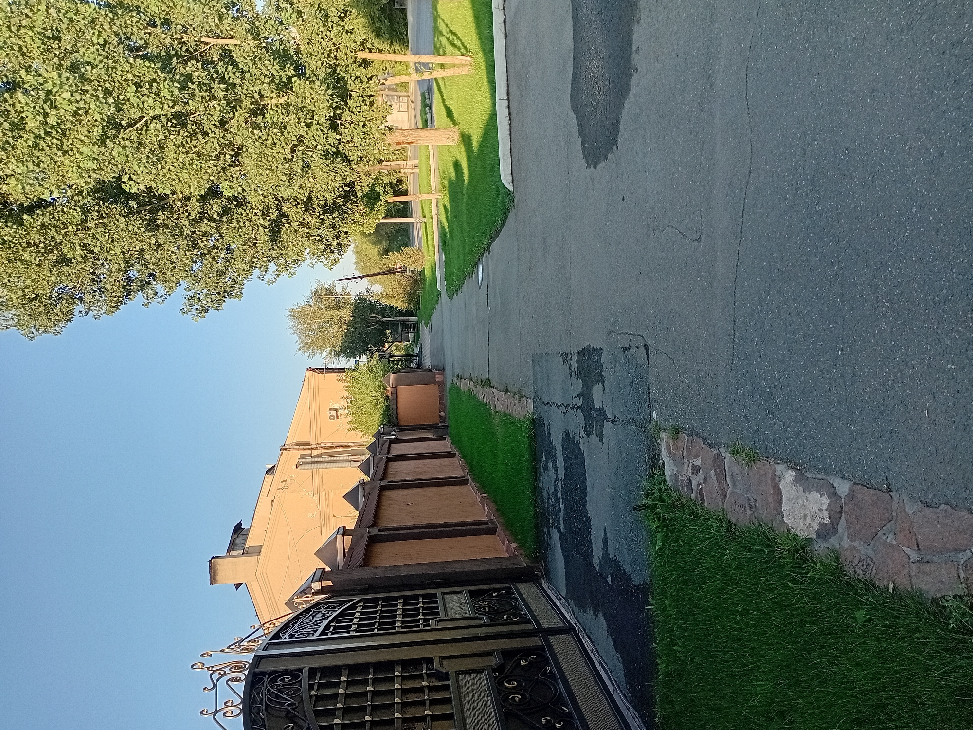 An outdoor, slightly elevated, full shot captures a paved sidewalk running alongside a decorative metal gate, a brown fence, and a two-story building. The building, painted a pale yellow, has a chimney and appears to be a residential or commercial structure. The fence has a darker brown trim and is interspersed with pointed decorative elements. A strip of neatly trimmed green grass runs along the base of the fence. The sidewalk itself is cracked and uneven, with some wet patches indicating recent rain or water spillage. To the right of the sidewalk, another stretch of manicured green grass is visible, bordered by a concrete curb. Several trees with dense green foliage are scattered throughout the scene, casting shadows on the grass and sidewalk. The sky is a clear, light blue. The location is Semey, Kazakhstan.
