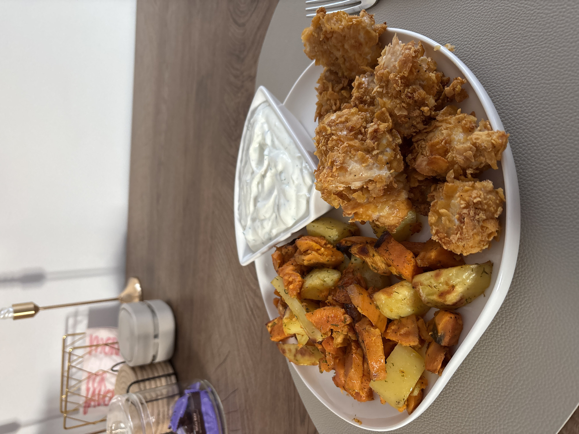 A white plate with a curved edge sits on a grey placemat. The plate contains a portion of roasted sweet potatoes and regular potatoes, seasoned with herbs, and a pile of breaded chicken pieces. A small white triangular dish filled with a creamy white sauce, possibly ranch dressing, is positioned on the plate behind the chicken. A silver fork rests to the right of the plate. In the background, on a wooden surface, are decorative items including a gold candlestick, a gold wire basket containing pink and white striped items, a small white cylindrical object, and a clear jar with a dark purple item. The scene appears to be indoors, likely in a dining area. The image is sharp and well-lit, with the food as the primary subject. The location is not discernible from the image itself, and therefore the claim that the image is located in Viernheim, Germany cannot be substantiated by visual evidence alone.
