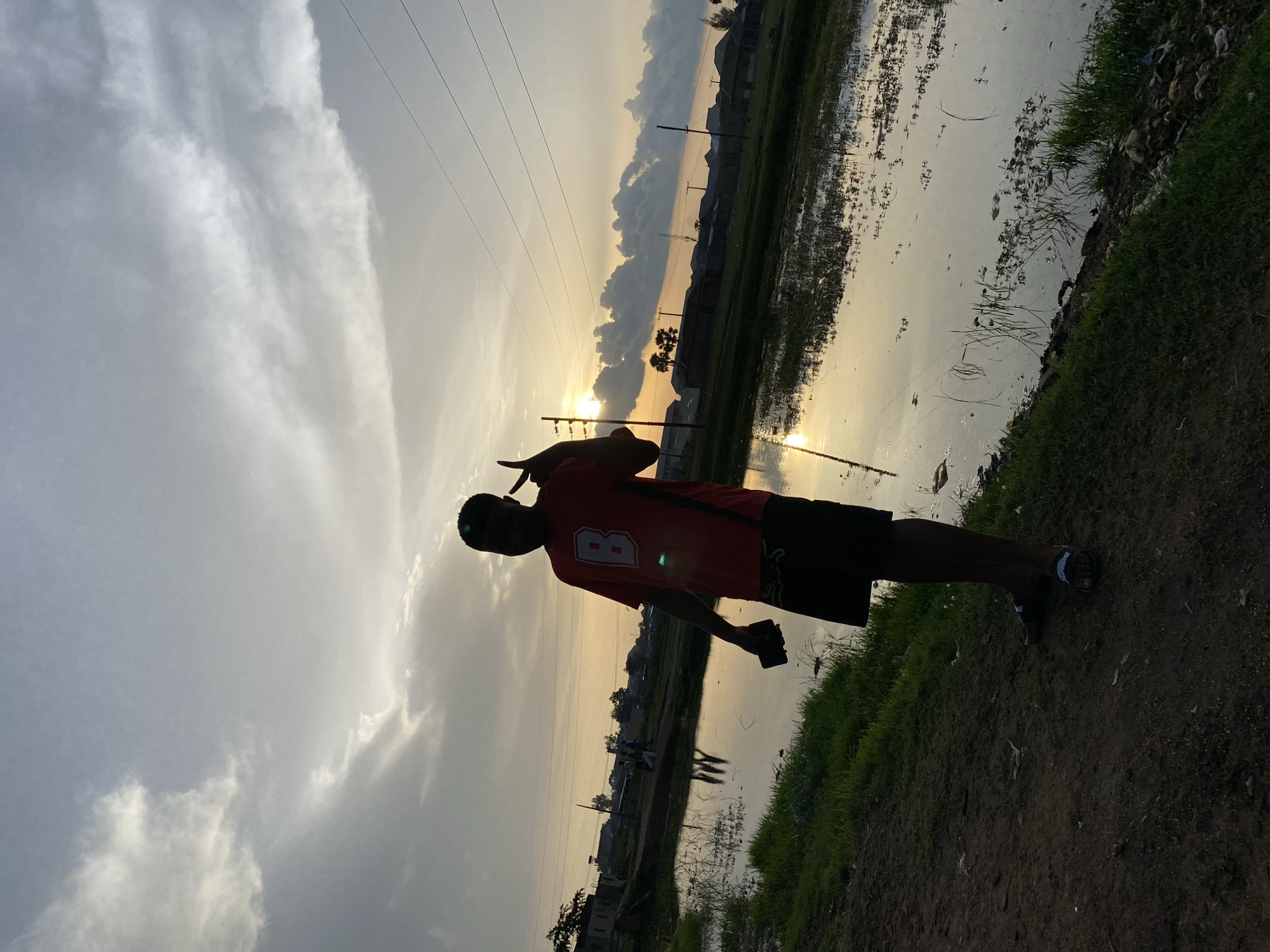 The image displays an outdoor scene at sunset in Bukuru, Nigeria. A young male, largely silhouetted, stands on a dirt path in the foreground, positioned to the left of the frame. He is wearing a red t-shirt with a white 'B' emblem, black shorts, and dark sandals. He holds a smartphone in his lowered left hand and displays a peace sign with his raised right hand.
The setting sun, a prominent bright disc, is low on the right horizon, casting a strong glare and an elongated reflection across a large, still body of water that occupies the central and right portions of the mid-ground. The sky features scattered clouds, displaying a warm gradient from yellow-orange near the horizon to light gray-blue overhead.
The foreground consists of dark earth transitioning to green grass bordering the water, which contains emergent aquatic vegetation. Multiple power lines and utility poles extend across the mid-ground. In the distance, numerous low-rise buildings with dark roofs are visible, interspersed with trees, including several palm trees. Two or three smaller silhouetted figures are discernible walking in the mid-ground on the left, near the water's edge.