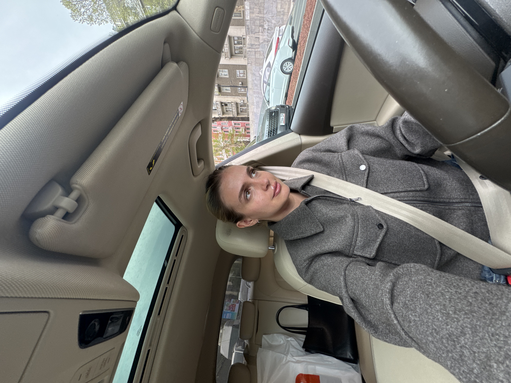 A woman with fair skin and light brown hair is seated in the driver's seat of a car. She is wearing a gray wool coat and a seatbelt. The car's interior is light beige. A black leather tote bag and some shopping bags are in the back seat. The car has a sunroof. Through the car window, buildings are visible. Based on the building architecture and vehicle license plates (2264-CP), the location is Tbilisi, Georgia.