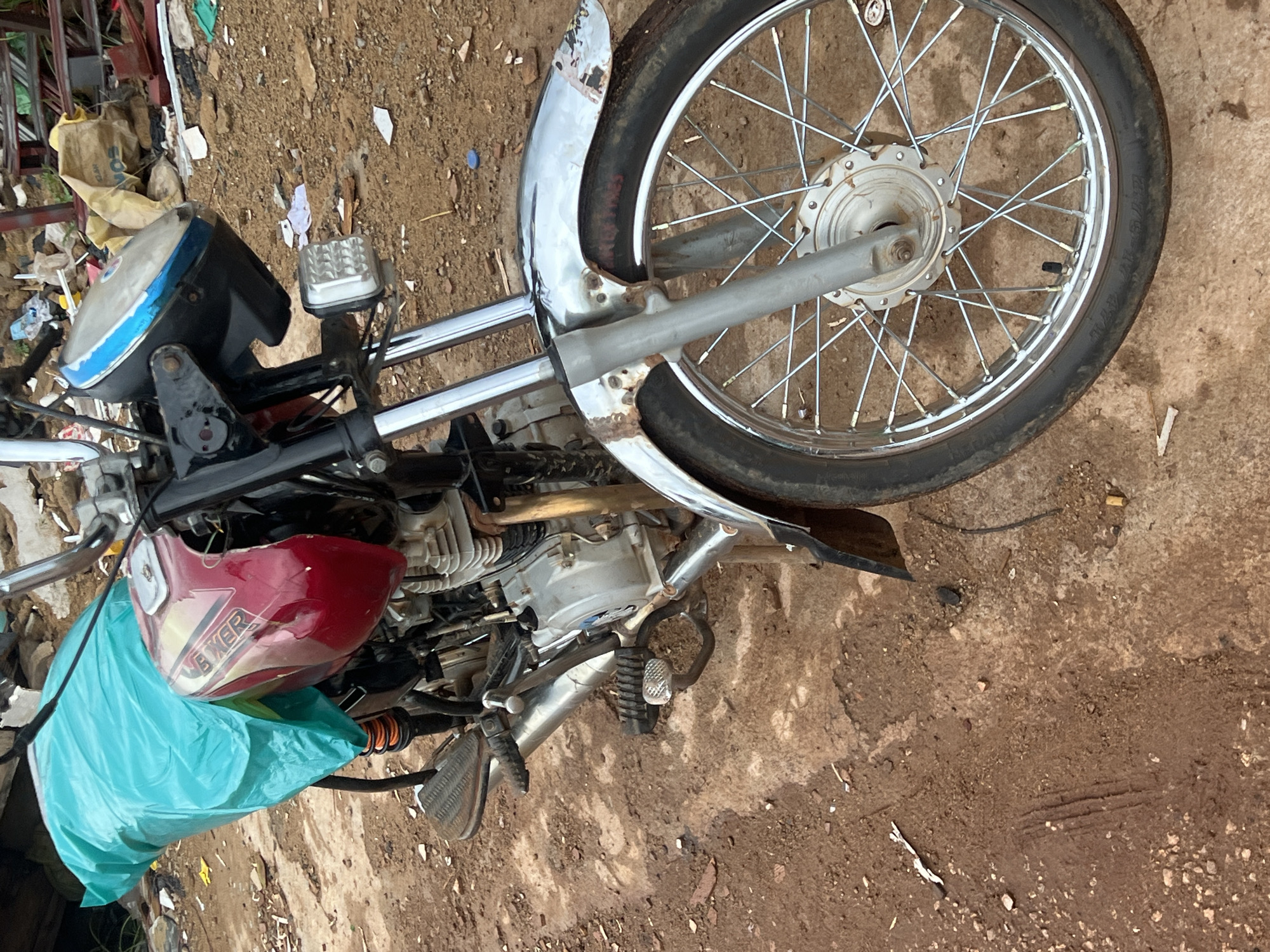 A close-up, eye-level shot shows the front portion of a red and silver motorcycle parked on a dirt ground, possibly in Durumi, Nigeria. The motorcycle has a turquoise bag strapped to the back. The front wheel is visible, as is the engine and part of the handlebars. The environment appears to be an outdoor, somewhat unkempt area with debris scattered on the ground. The lighting suggests it is daytime.
