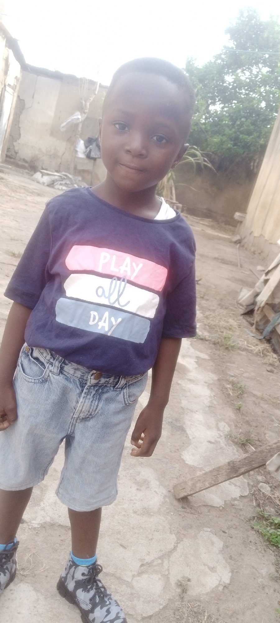 A young boy stands outdoors in Sabon Tasha, Nigeria. He wears a navy blue t-shirt with horizontal pink, white, and blue stripes and the words "PLAY all DAY", denim shorts, blue socks, and camouflage high-top sneakers. The environment appears to be a residential area with unfinished buildings and foliage visible in the background. The ground is a mix of concrete and dirt.