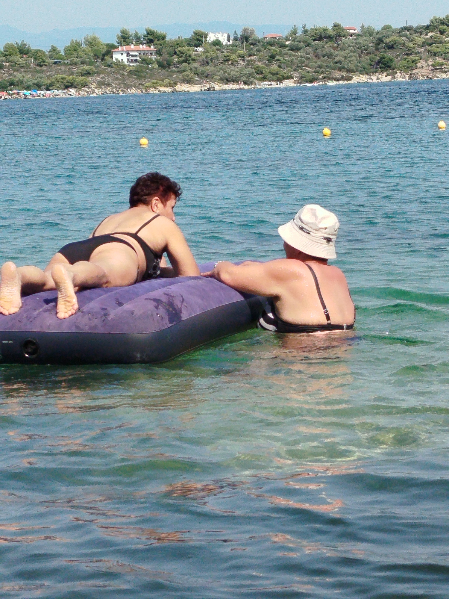 Two women are floating on an inflatable mattress in the Aegean Sea off the coast of Sithonia Municipal Unit, Greece. The woman in the foreground, on the left, is lying face down with her legs extended. She is wearing a black bikini. The woman on the right is sitting with her back to the camera, wearing a white bucket hat and a black and white striped bikini. The water is clear and turquoise, with gentle waves. In the background, a coastline with lush green hills and white buildings can be seen, indicating a populated area. Yellow buoys are scattered in the sea. The overall scene depicts a peaceful day at the beach.