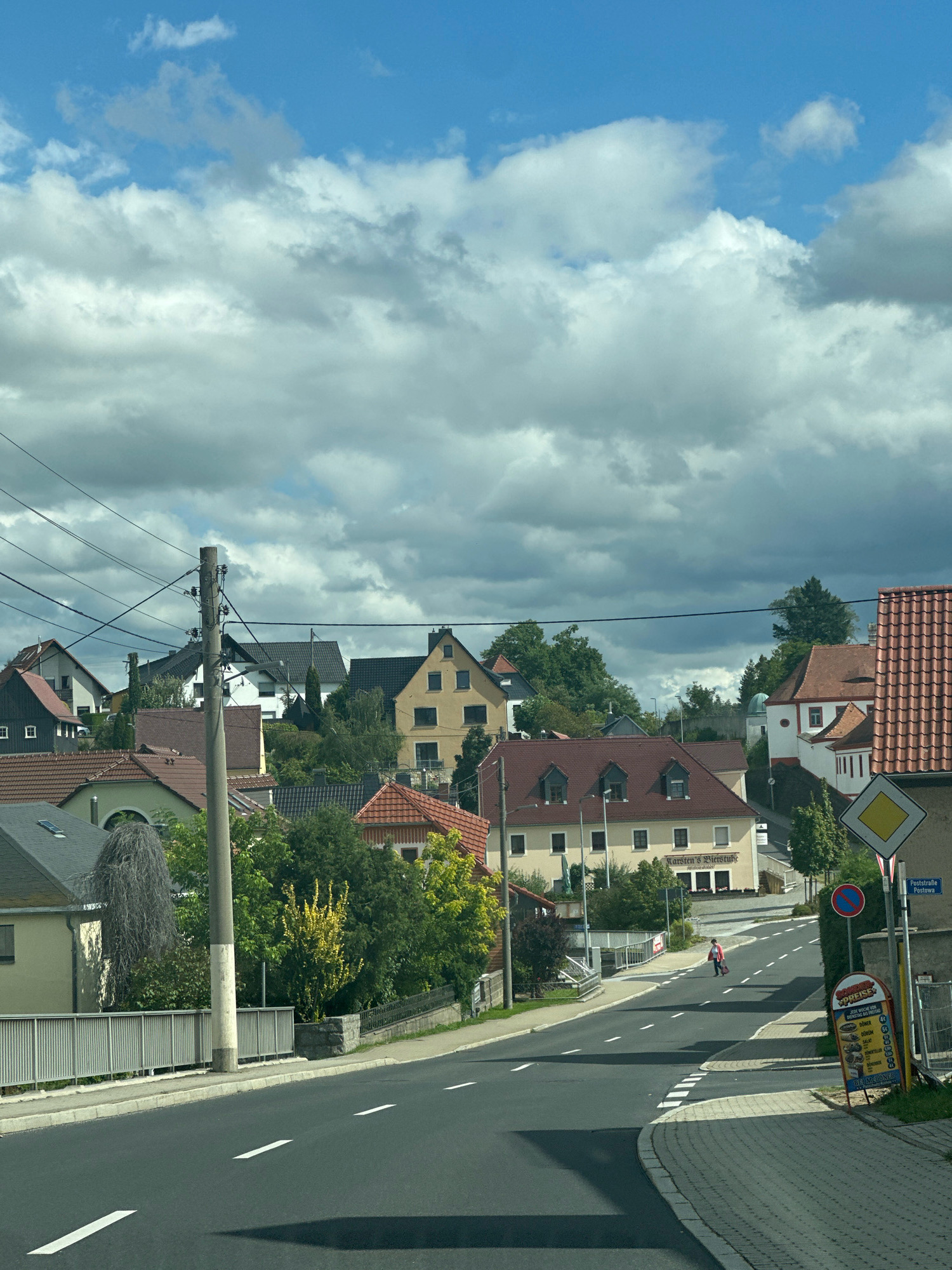 The image depicts a street scene in Panschwitz-Kuckau - Pančicy-Kukow, Germany. The foreground is dominated by a paved road with white lane markings. To the left, a grey metal fence with vertical bars runs alongside the road, with a concrete sidewalk behind it. A prominent concrete utility pole with electrical wires stretches across the upper left portion of the image.
Beyond the fence and road, a cluster of houses with red-tiled roofs and neutral-colored facades are visible, interspersed with green trees. The houses exhibit varying architectural styles, with some featuring dormer windows. A street sign with "Poststraße Postowa" written on it is located on the right side of the road, along with a yellow diamond-shaped yield sign and a circular red and blue sign. Below these, a vertical sign displays food items and prices in German.
A single person, wearing a pink top and dark bottoms, is walking across the street in the mid-ground, carrying a dark bag. The sky above is a mixture of bright blue and heavy grey clouds. The overall impression is of a residential area on a cloudy day.