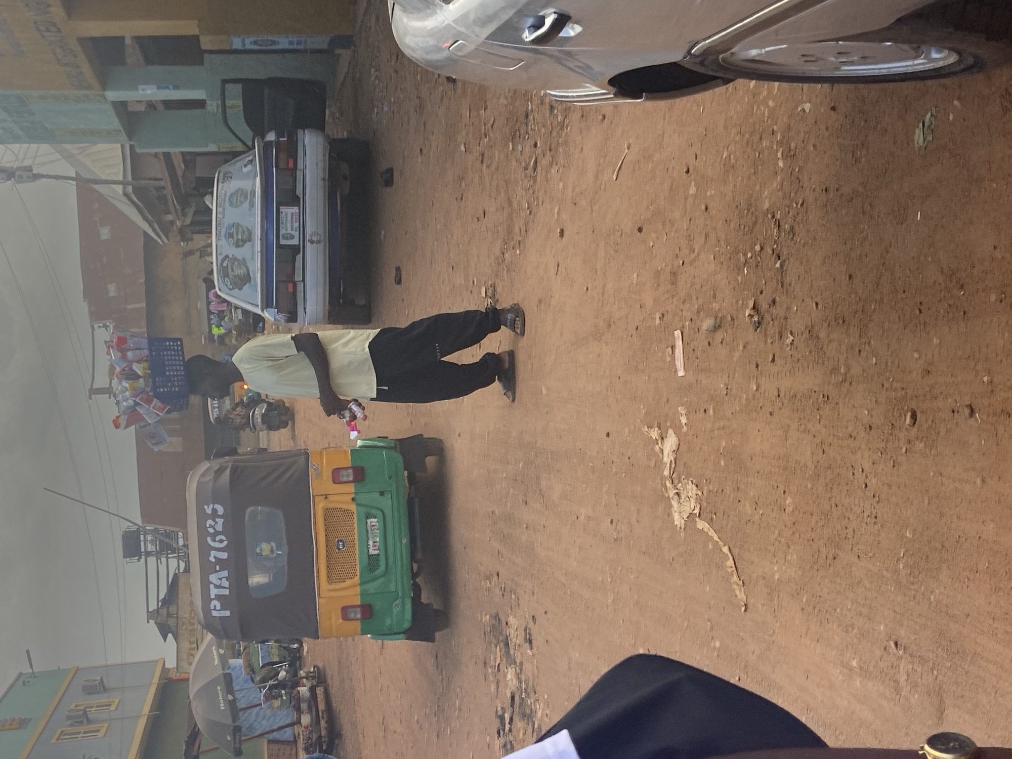 In Bukuru, Nigeria, a man stands in the middle of a dirt road, balancing a basket of goods on his head. He wears a light yellow t-shirt and black pants. To his left, a green and yellow auto-rickshaw, with license plate PTA-7625, is parked. To his right, a white car with tinted windows is parked, its driver's side door open. The car's rear window displays three portraits of men and the text "TINUBU & MUJTAMA 2023". Buildings line the street, some appearing commercial with awnings and signs. The sky is overcast, suggesting a cloudy day. The ground is a reddish-brown dirt, littered with small stones and debris.