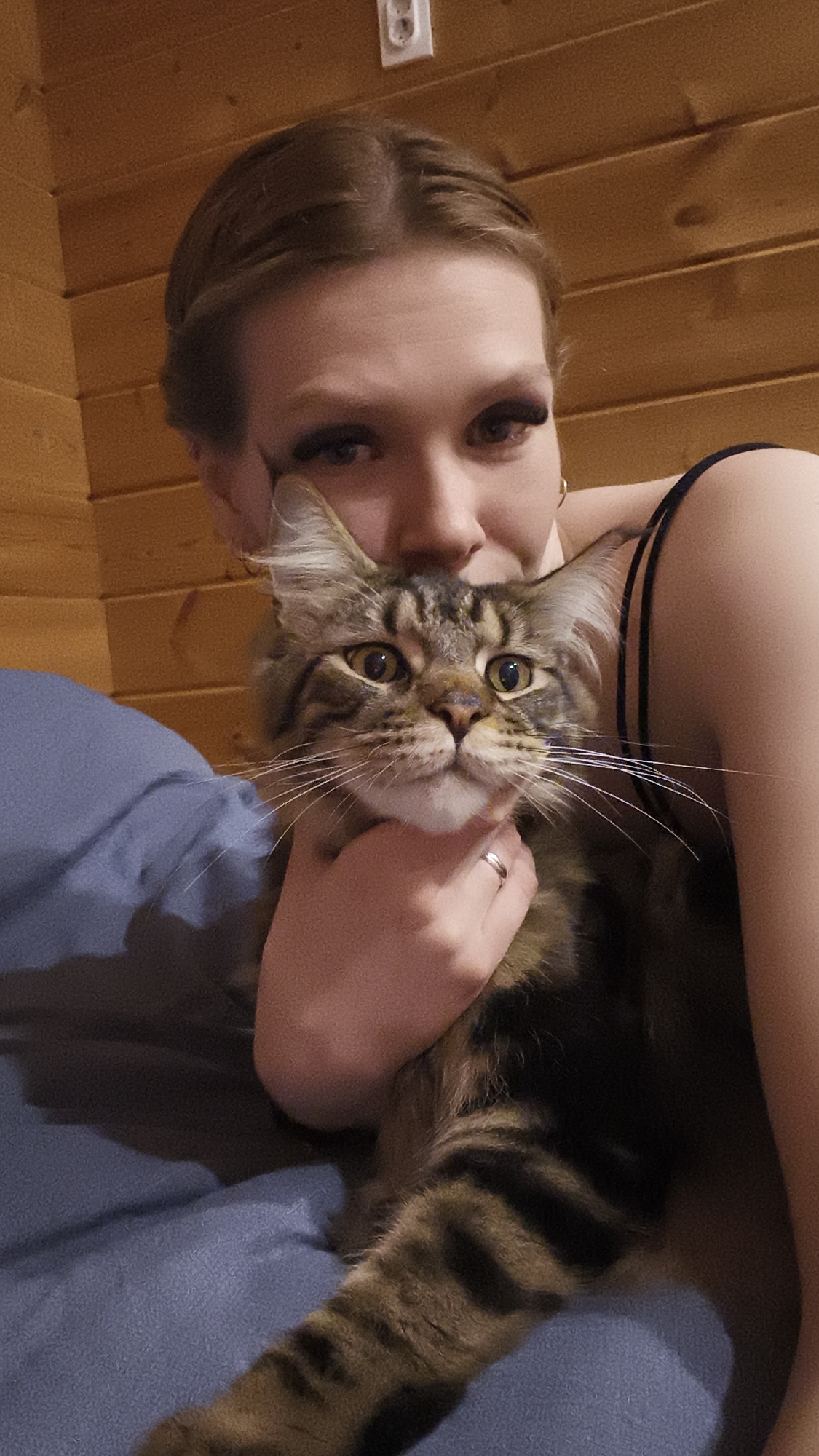 A woman with light brown hair, possibly in her late teens or early twenties, is holding a Maine Coon cat. The cat is brown tabby, and the woman is holding it with her right hand, which has a ring on her ring finger. The background is a wood-paneled wall with an electrical outlet. A blue surface is visible beneath the cat. There are no explicit references to the location being Русский Вожой, Russia, in the image.